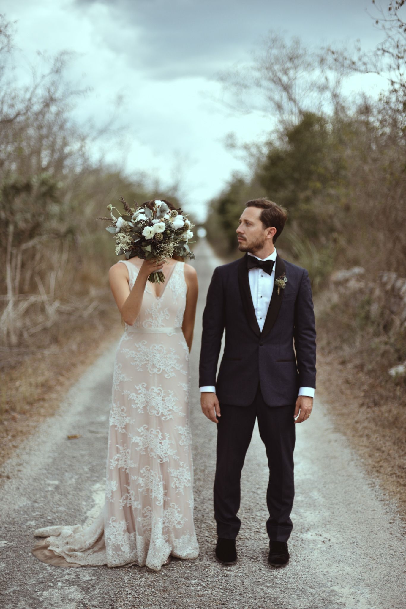 A couple in wedding attire stands on a rustic dirt path, with the bride holding a bouquet in front of her face.