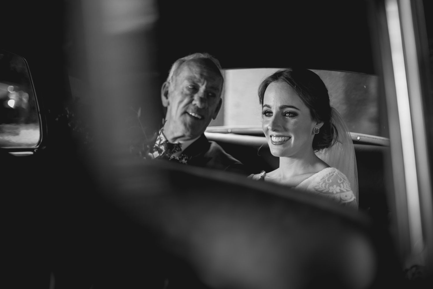 Black and white photo of two people smiling while sitting in a car together, viewed through the window reflection.