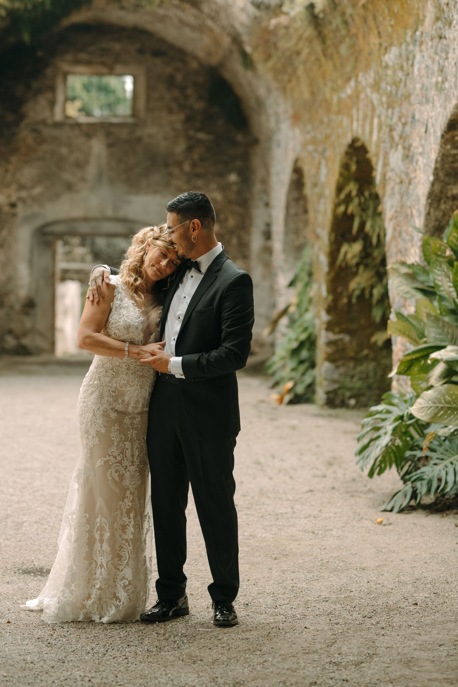 Intimate wedding moment captured in arched stone passageway with green plants.