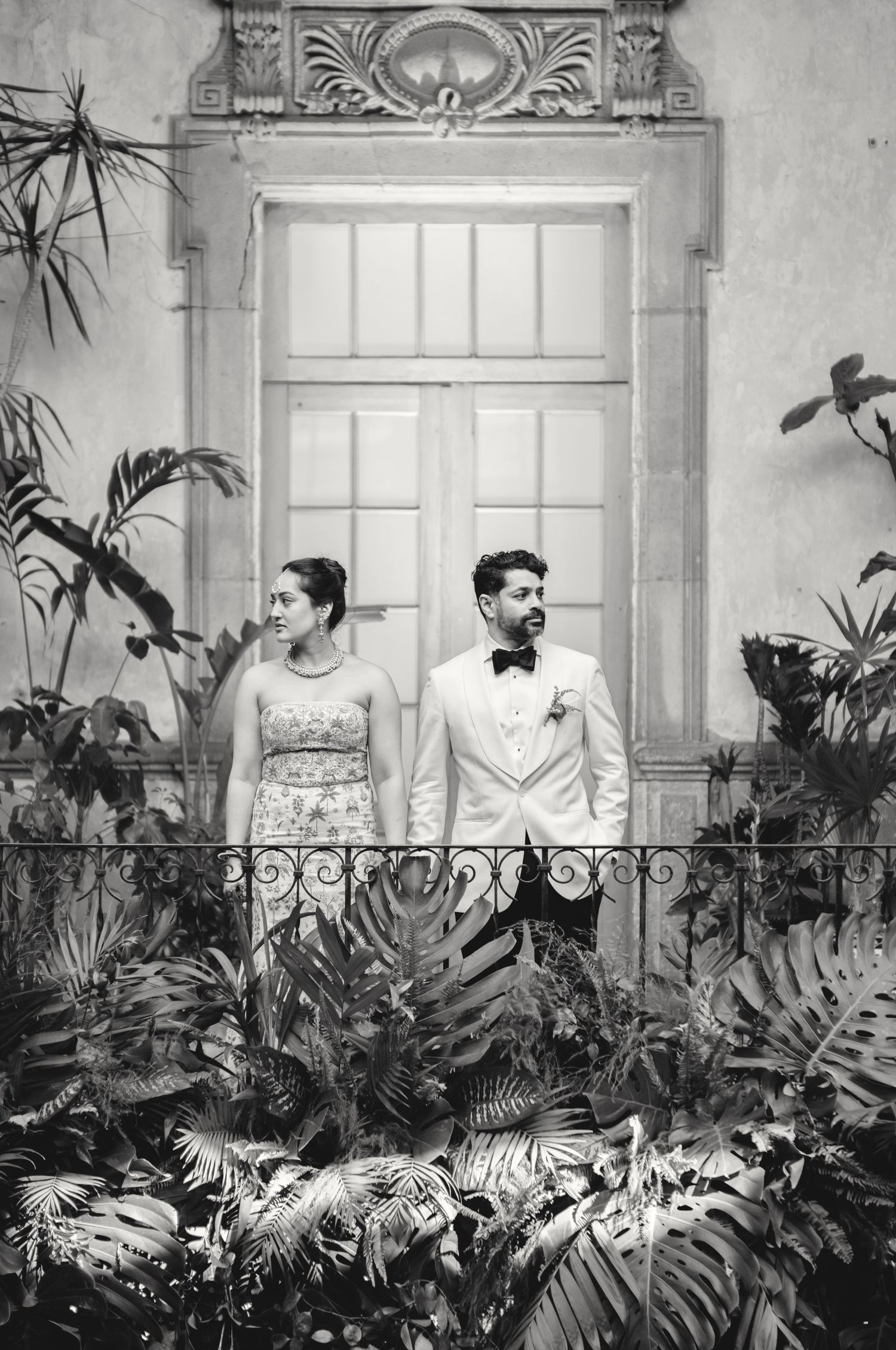 A couple poses in formal attire among tropical plants in a classic black and white portrait.
