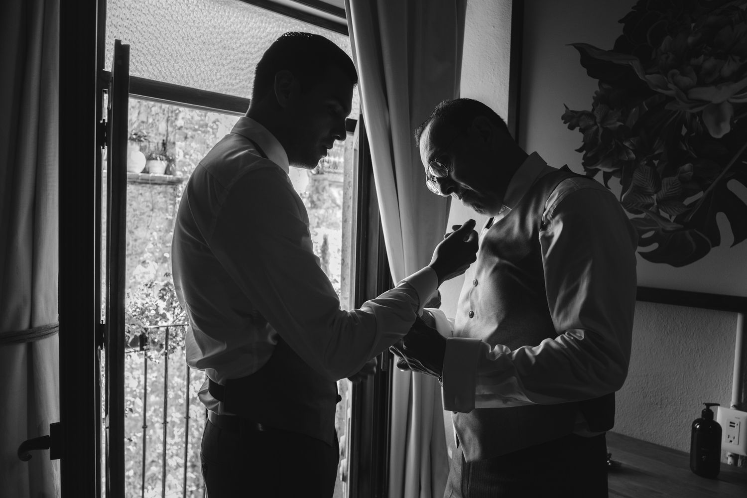 Black and white photo of two people helping with formal attire preparations near window.