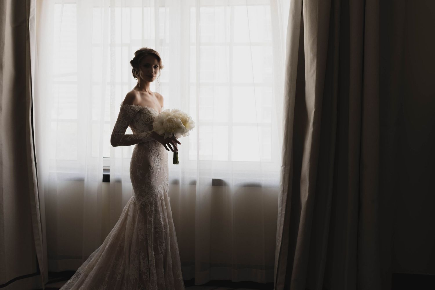 Silhouette of a bridal figure holding a bouquet against backlit window.