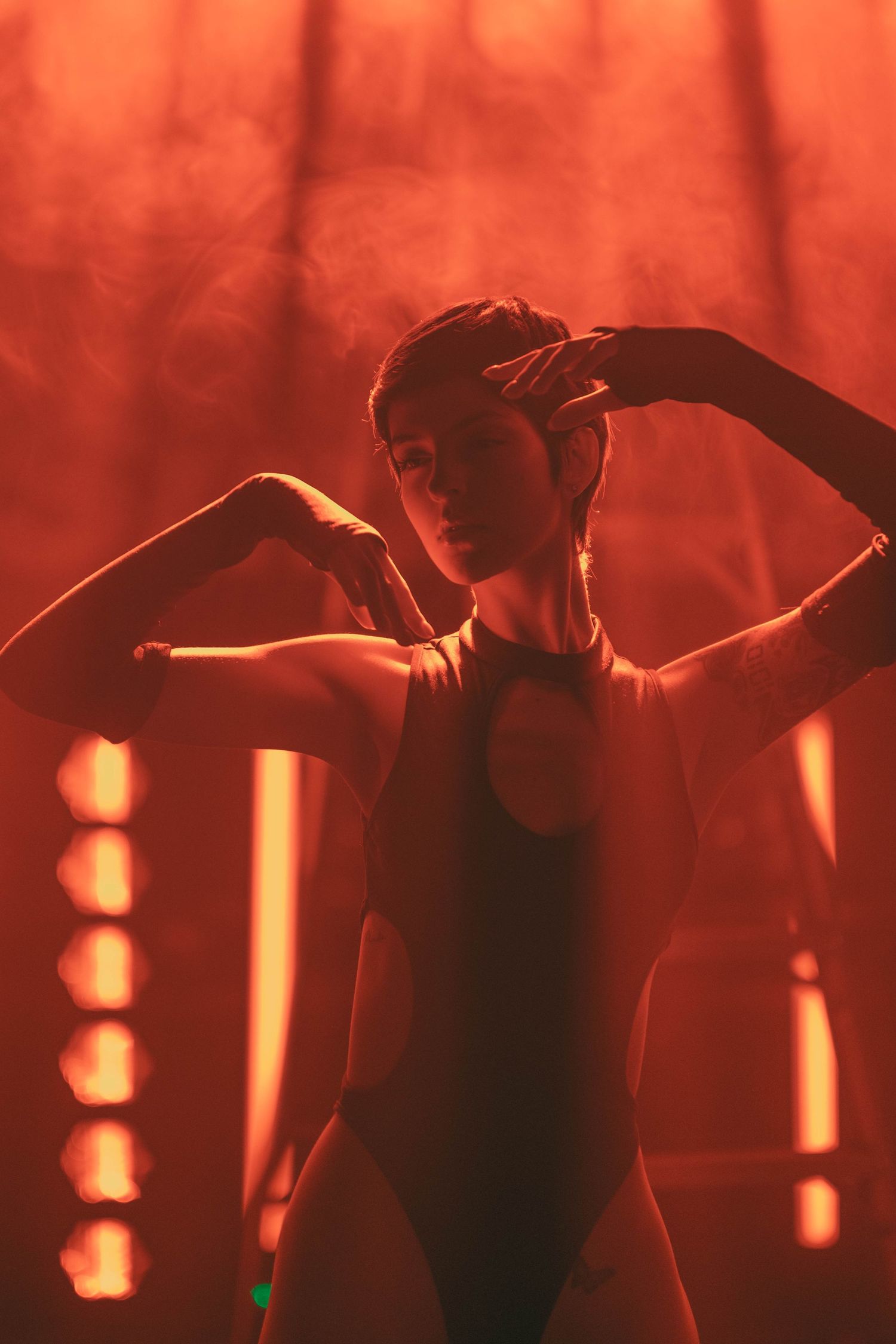 A dancer in a black leotard performs against a dramatic red-lit background with vertical lighting elements.