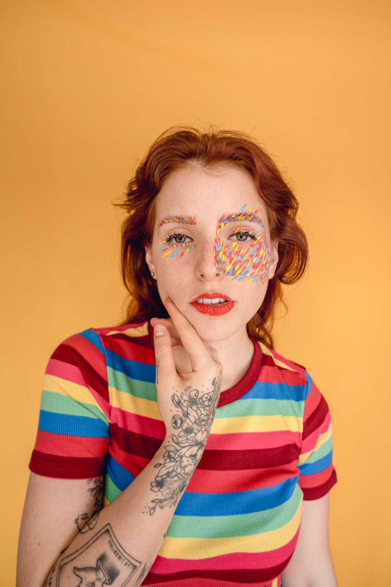 Portrait in rainbow striped shirt with face art and visible tattoos against orange background.