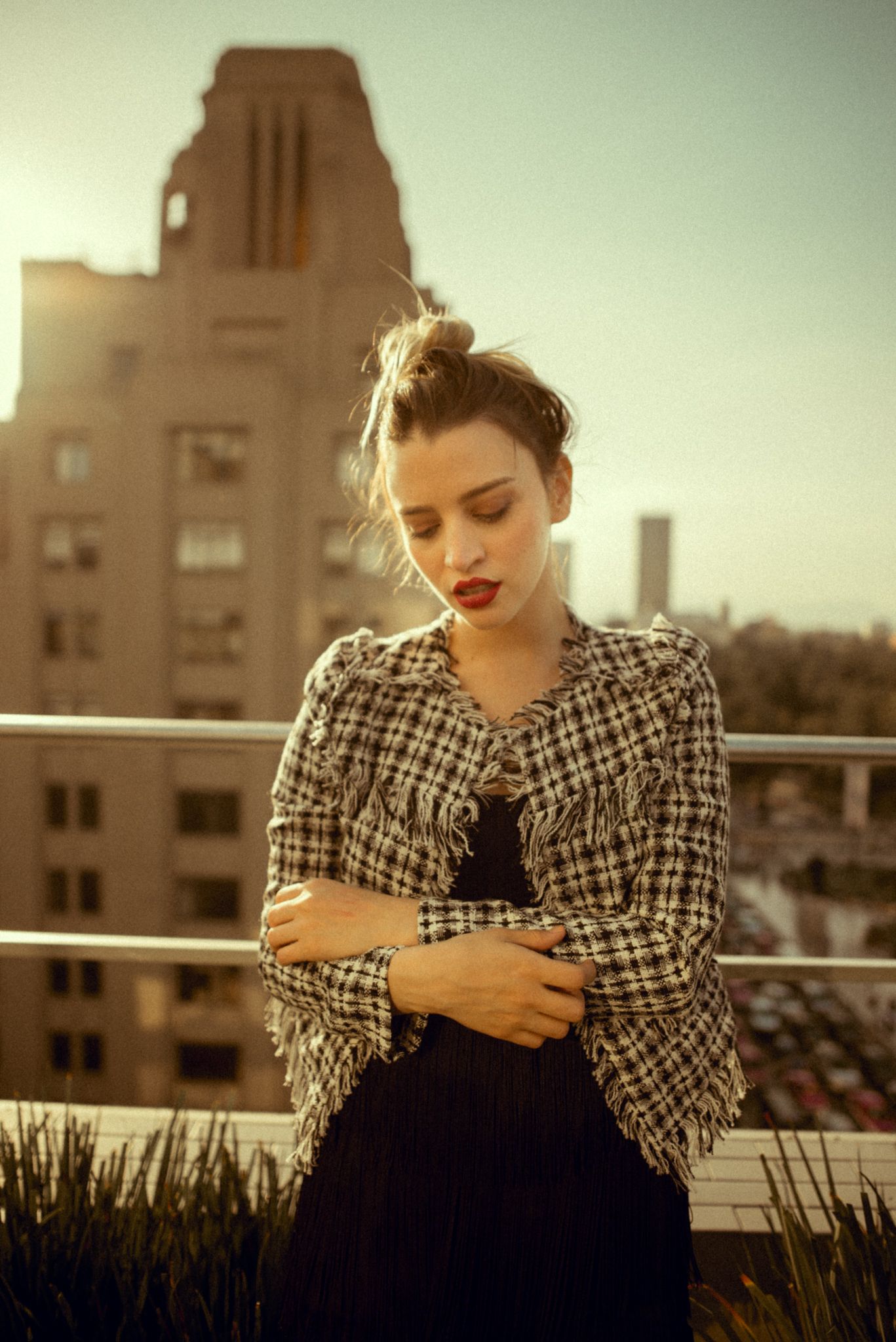 Fashion portrait on rooftop with plaid jacket and black dress against urban backdrop at sunset.
