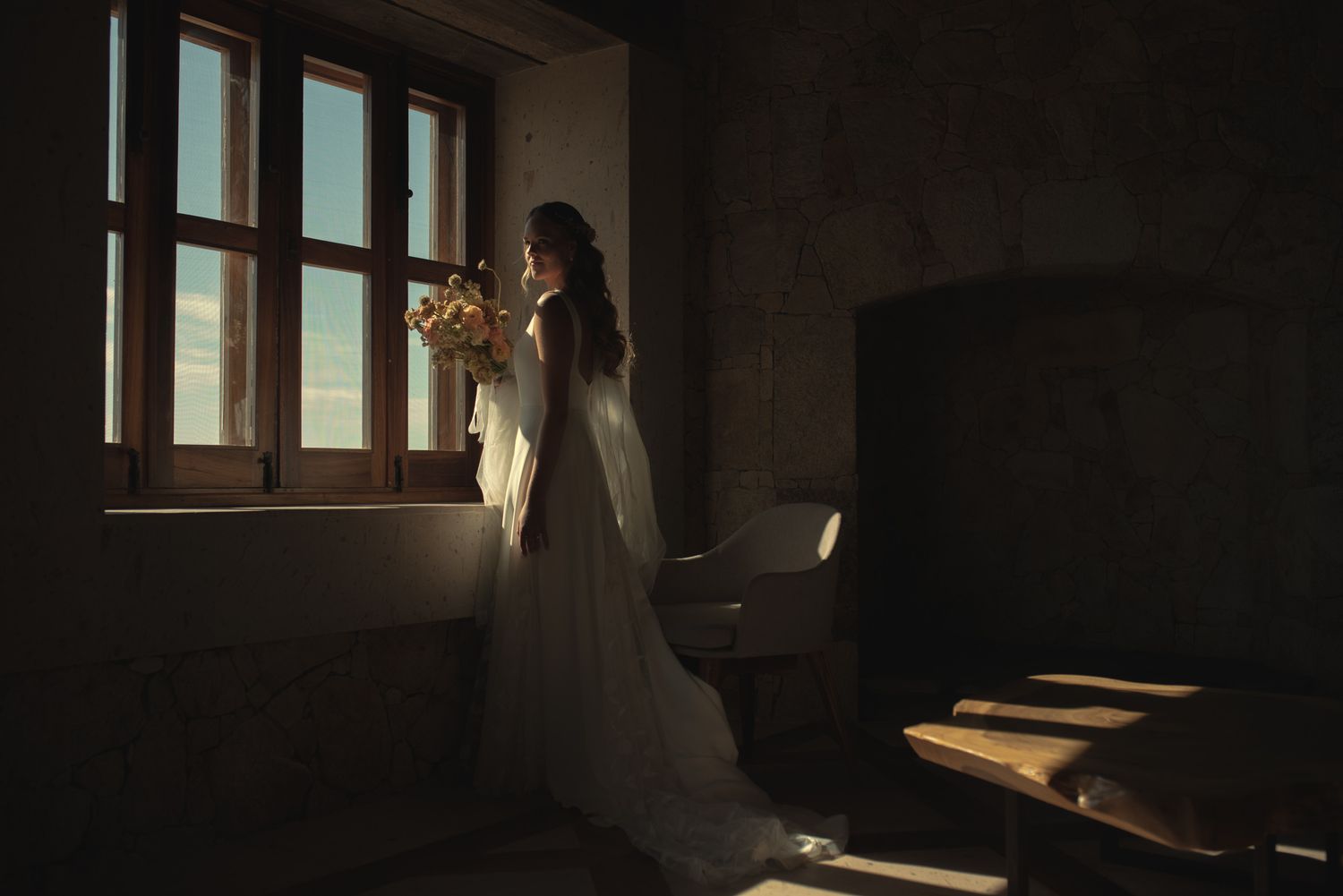 A silhouetted figure in a wedding dress stands by a window in a dark moody room holding a bouquet.