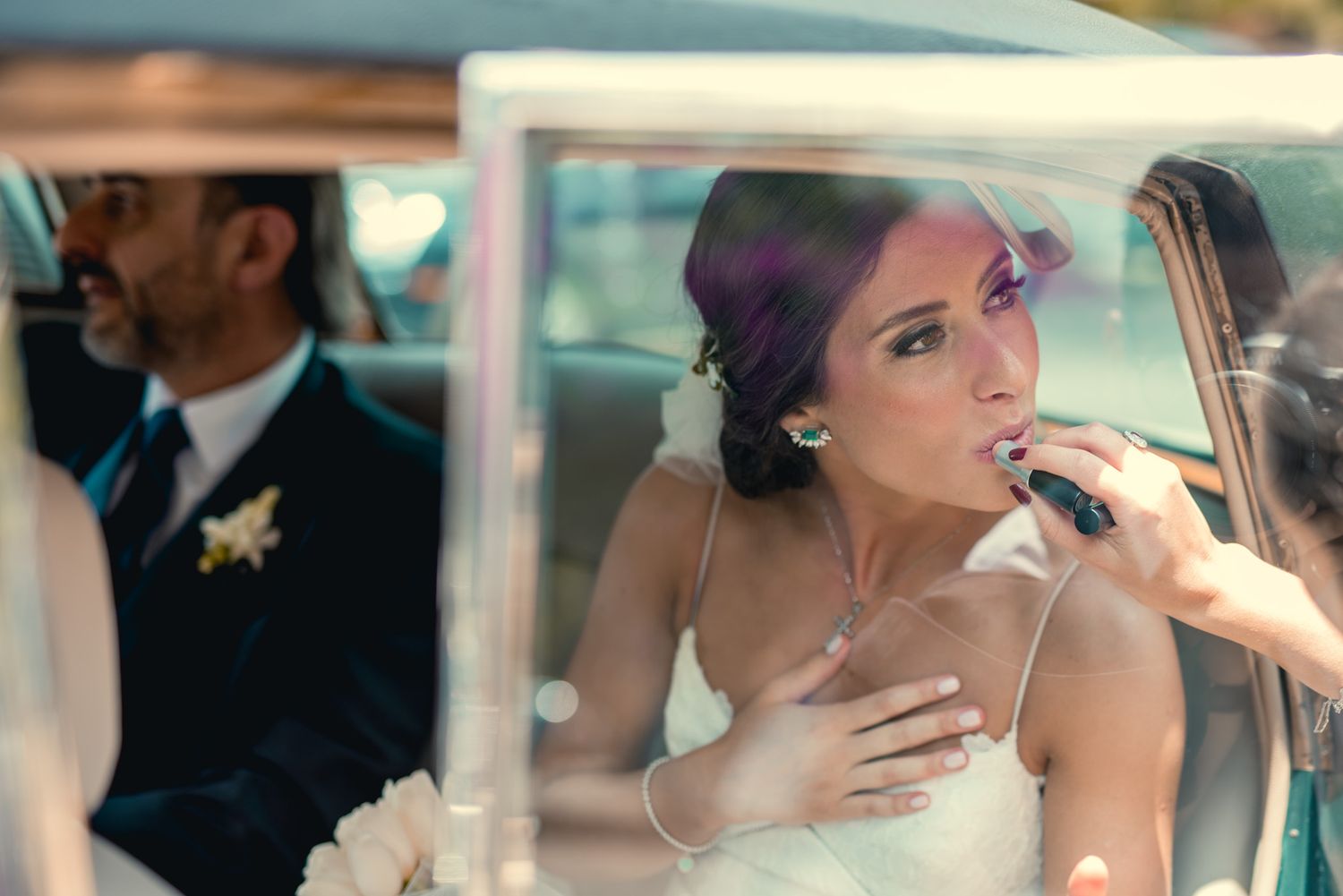 A person in a white wedding dress sits in a vintage car while receiving final makeup touch-ups.
