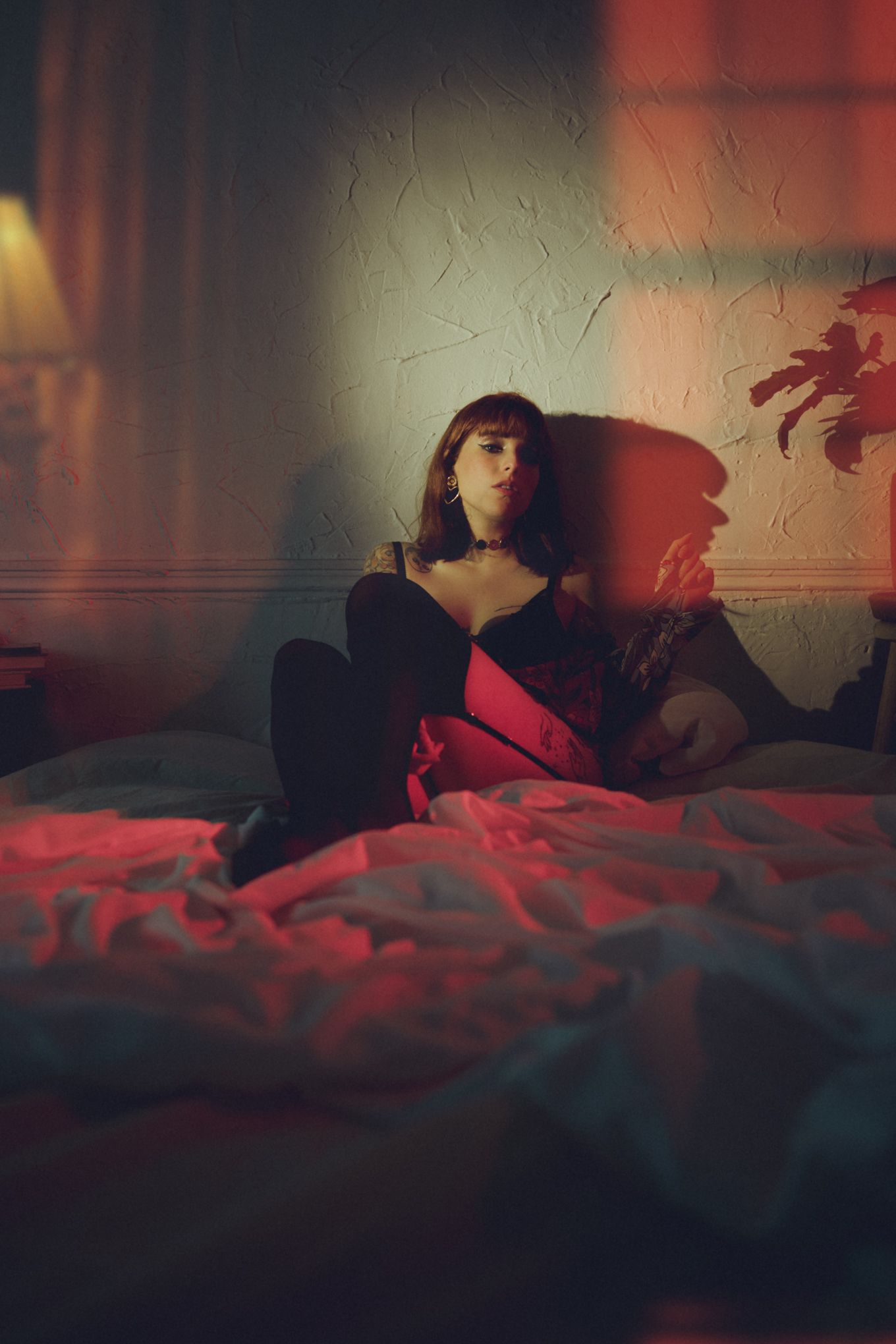 Figure reclining on bed in moody red-lit room creating atmospheric portrait.