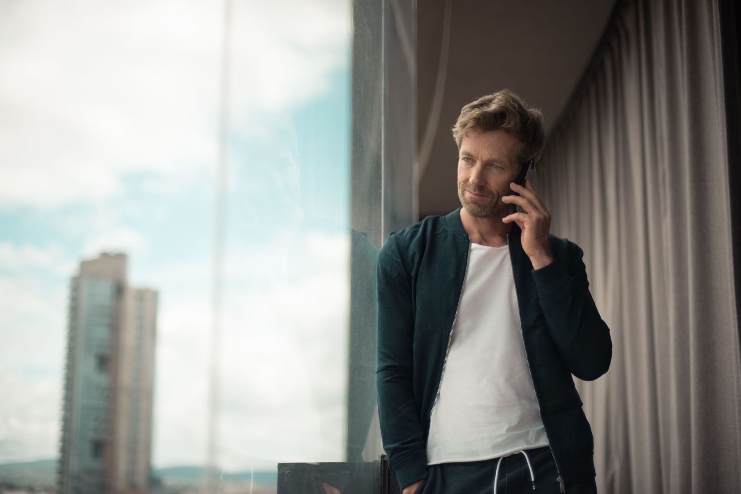 Person in casual attire talking on phone by window in modern high-rise building.