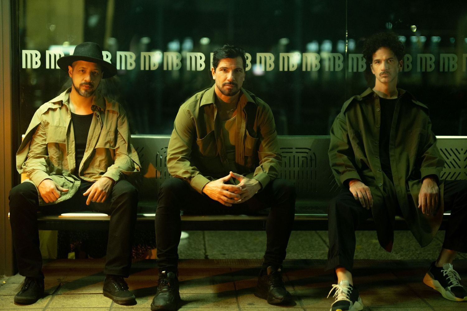 Three people sit on a bench against a dark wall with illuminated letters, creating a moody atmosphere.