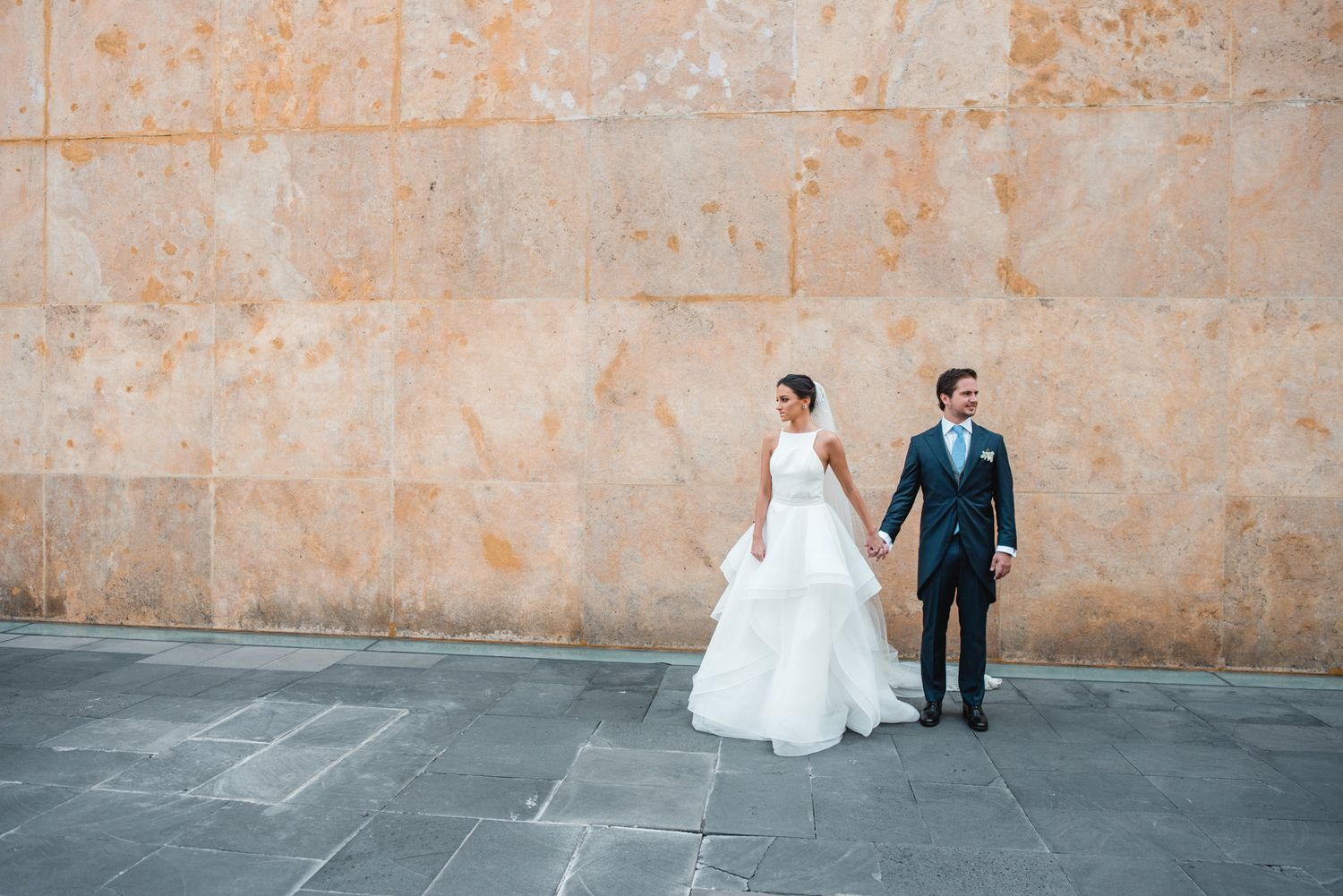 Bride and groom holding hands against a stone wall backdrop.