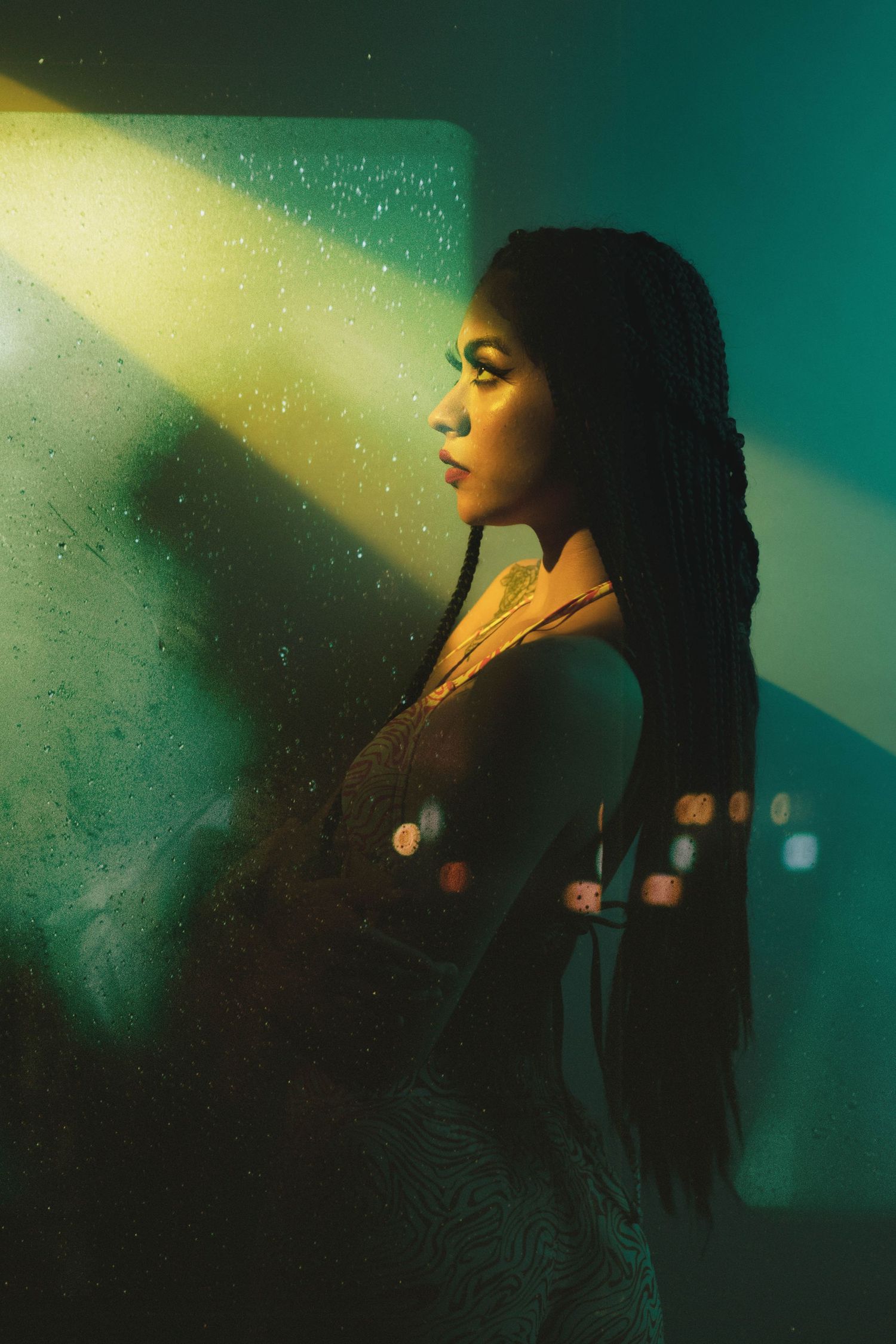 Artistic portrait in profile view against turquoise and yellow lighting through textured glass creating a dreamy atmosphere.