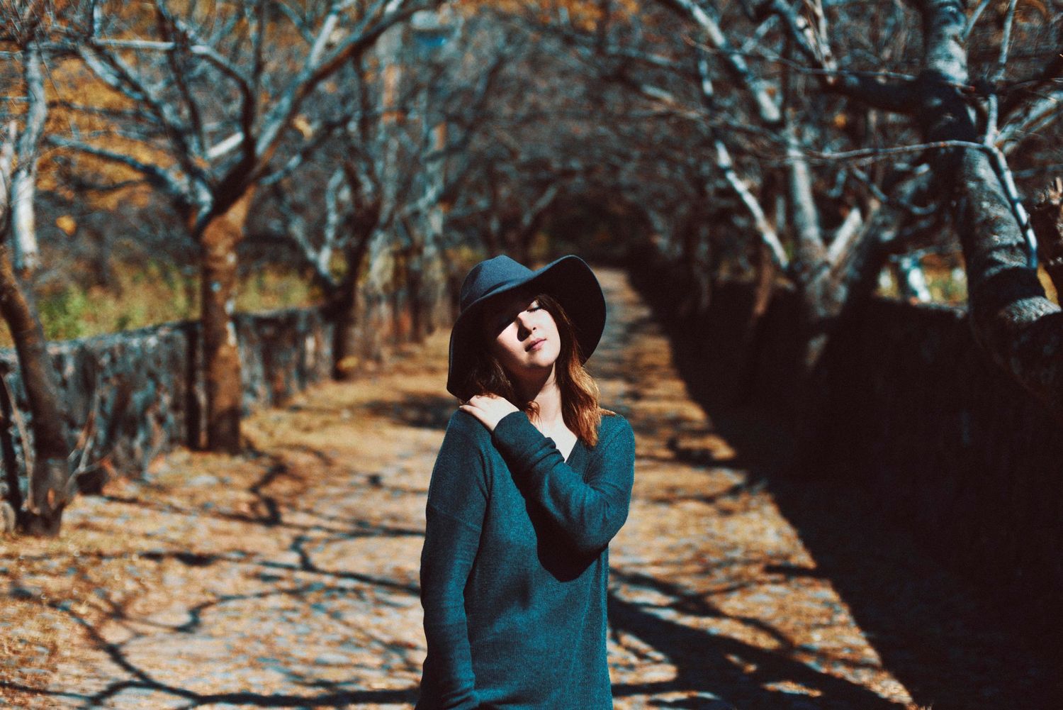 Autumn portrait of person in teal sweater and hat standing among bare trees.