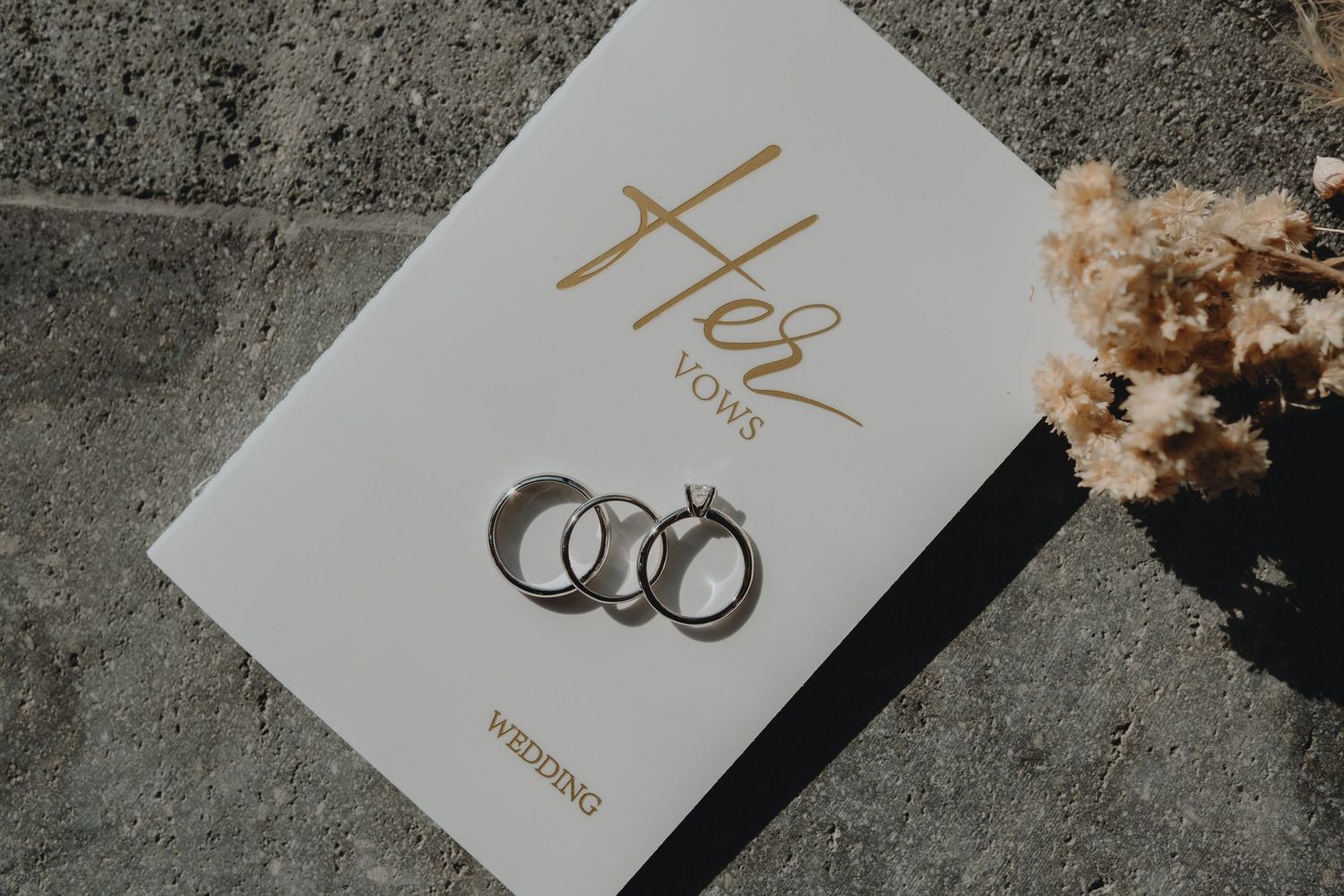 Wedding rings and vow book photographed on concrete surface with natural lighting.