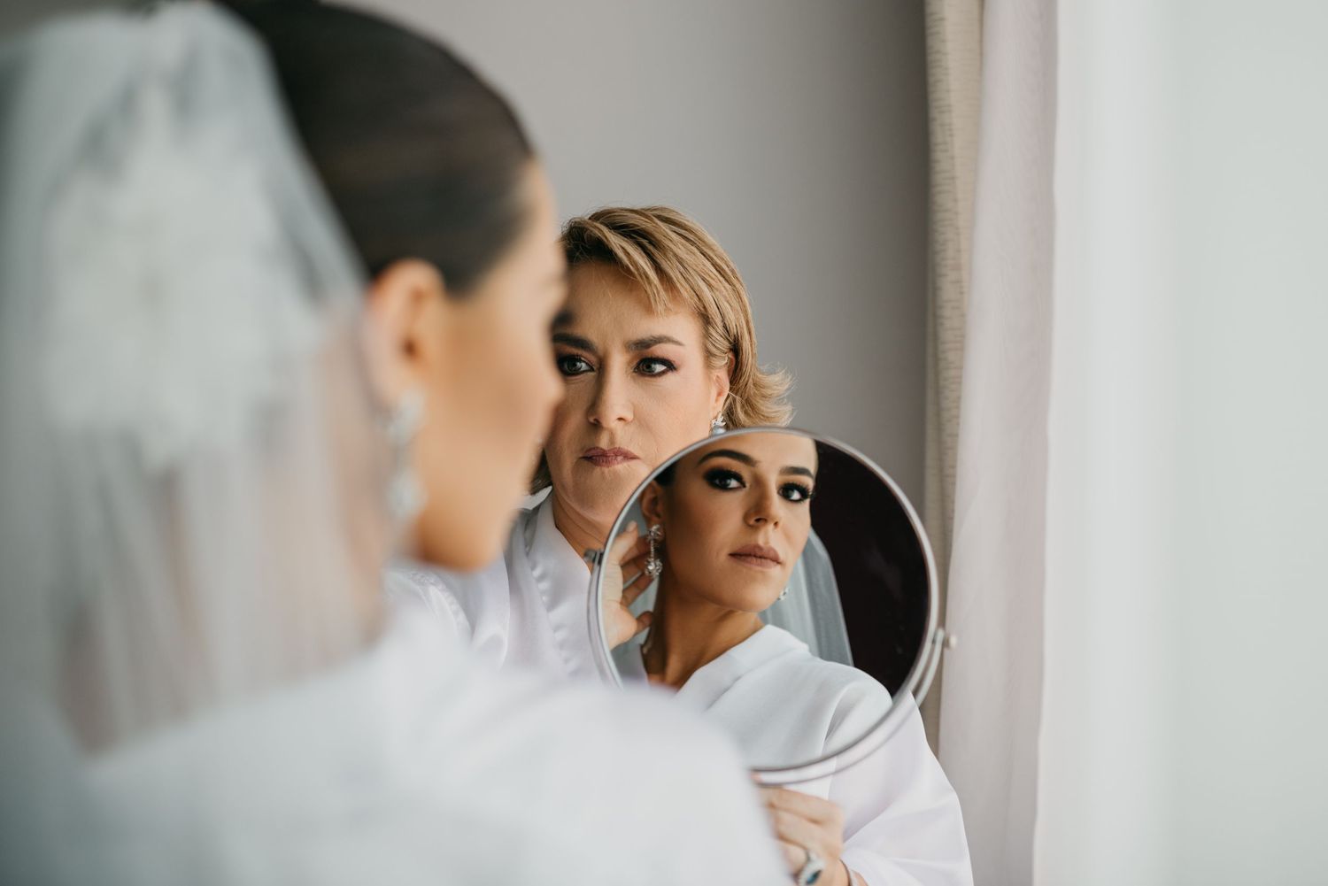 Two hairstylists work on preparing a bride's hair and makeup in front of a mirror during wedding preparations.