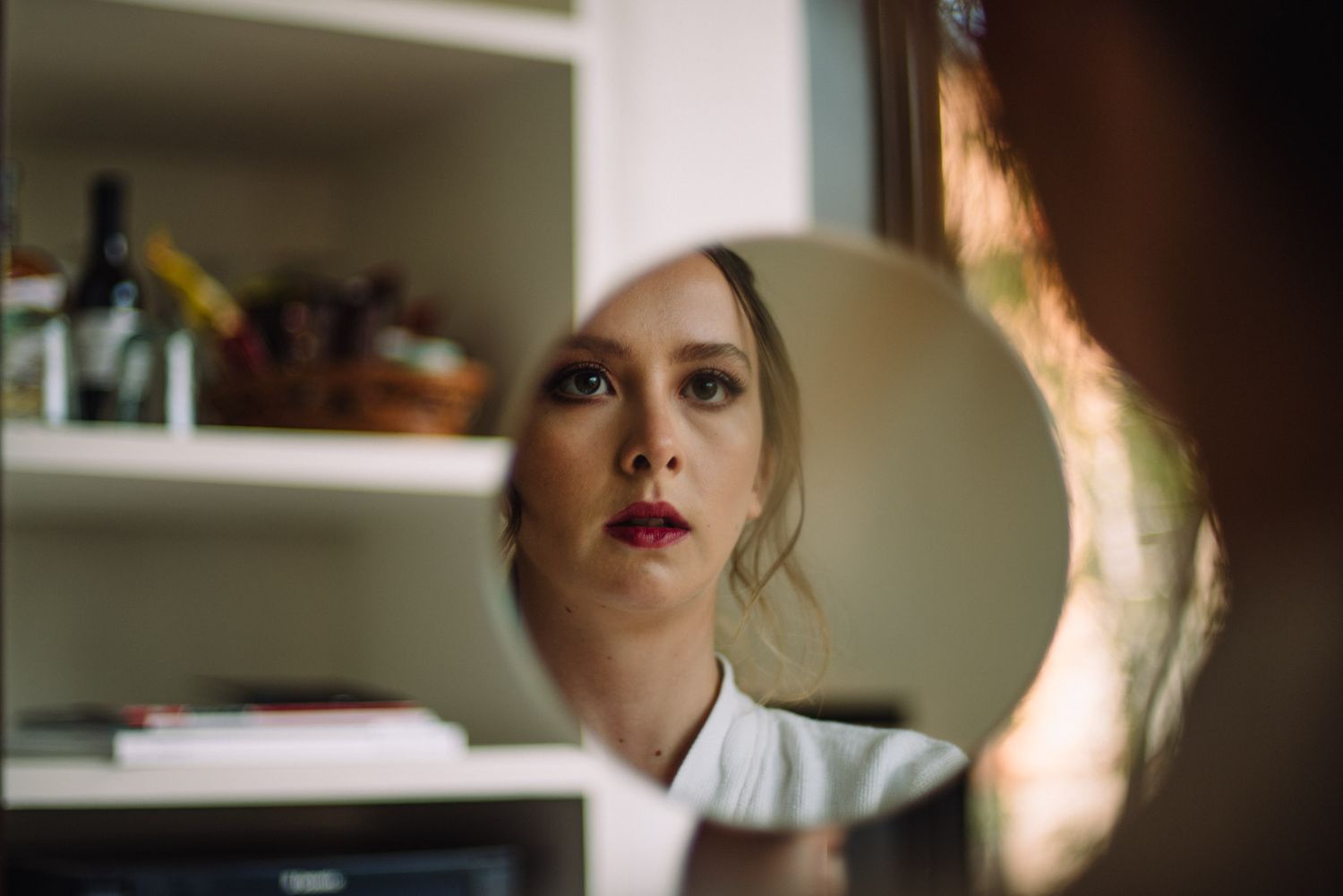 A reflection in a circular mirror shows a figure with red lipstick against shelving in the background.