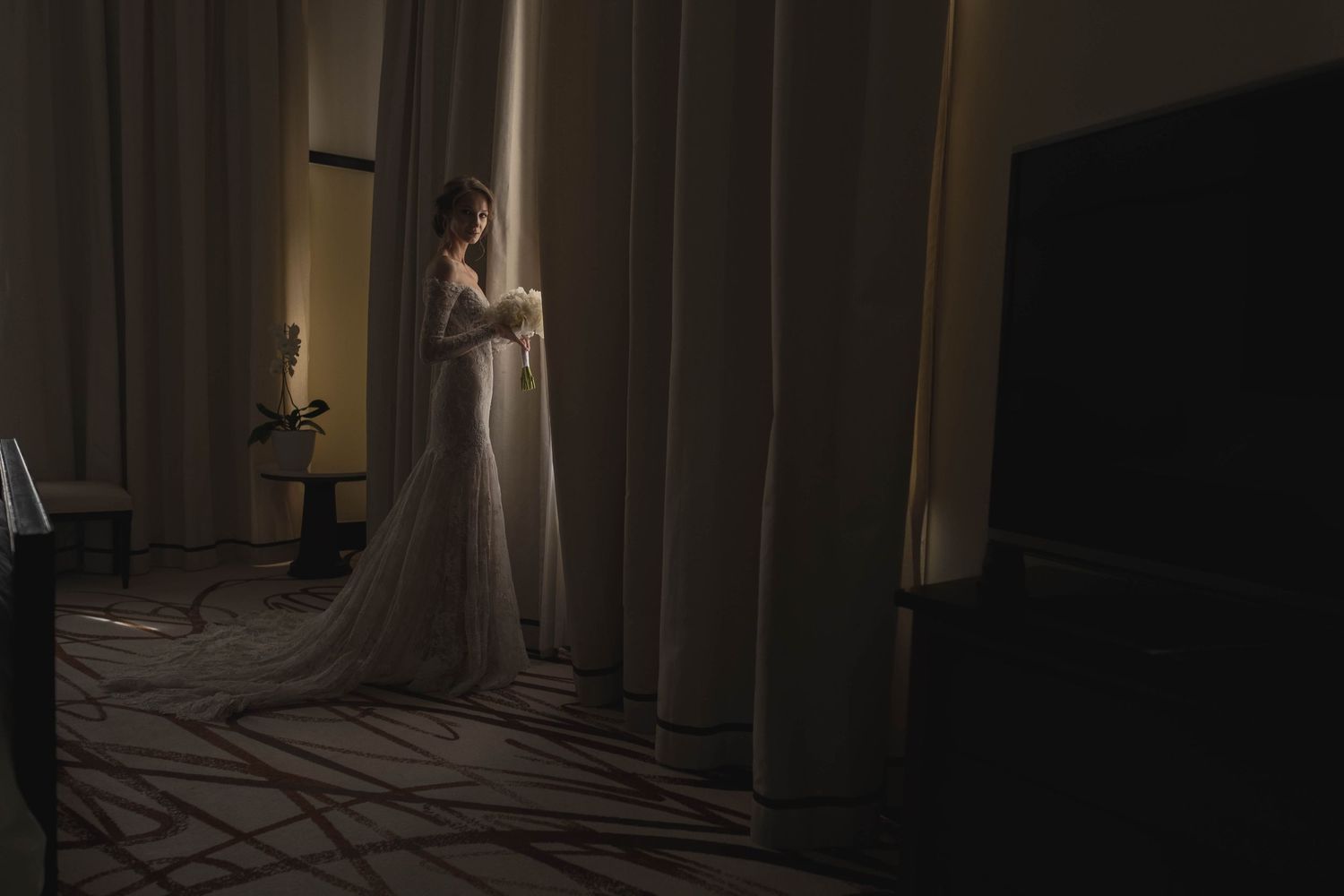 A bridal figure stands dramatically lit between curtains in a dim room.