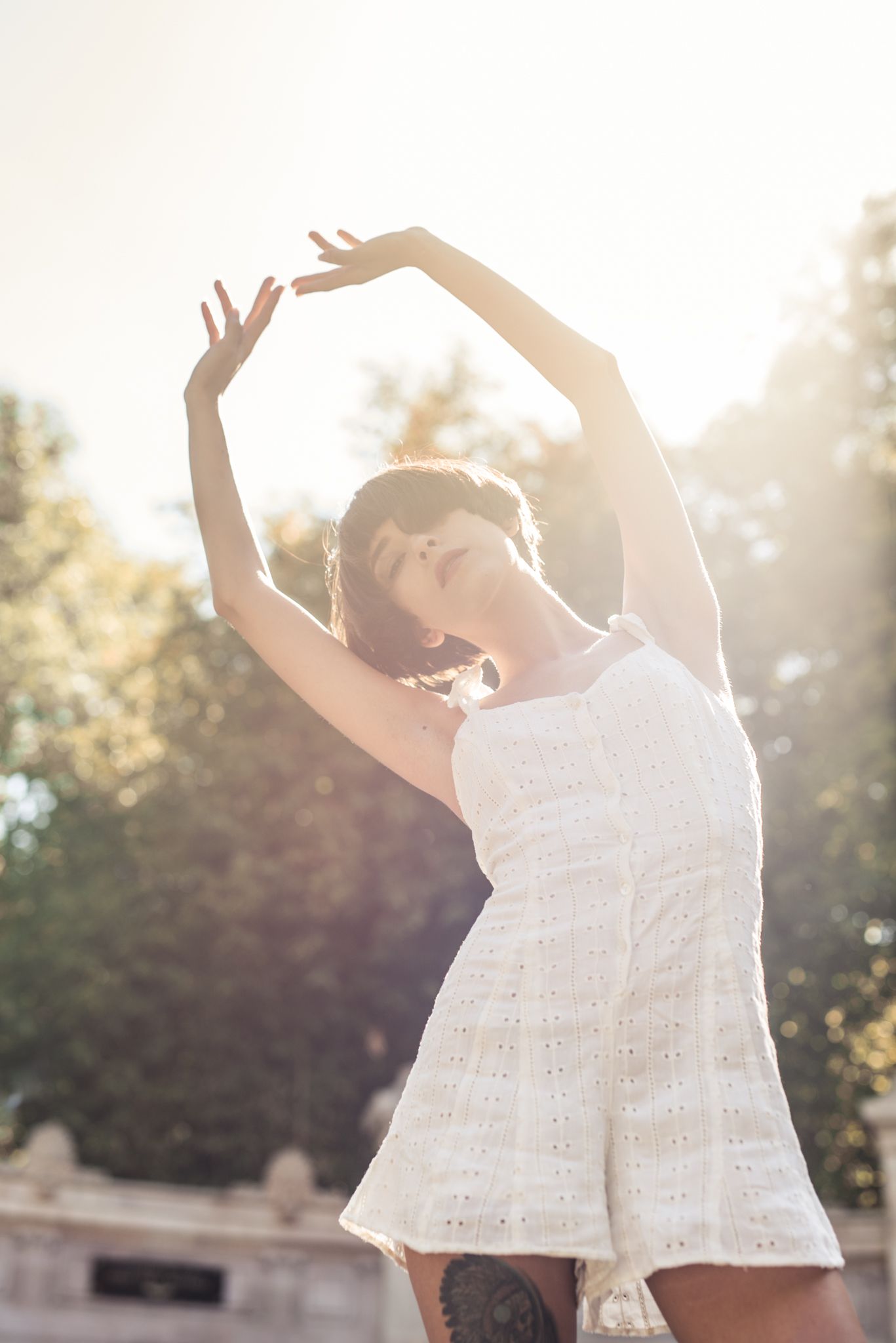Backlit figure in white dress dancing with raised arms in sunlight.