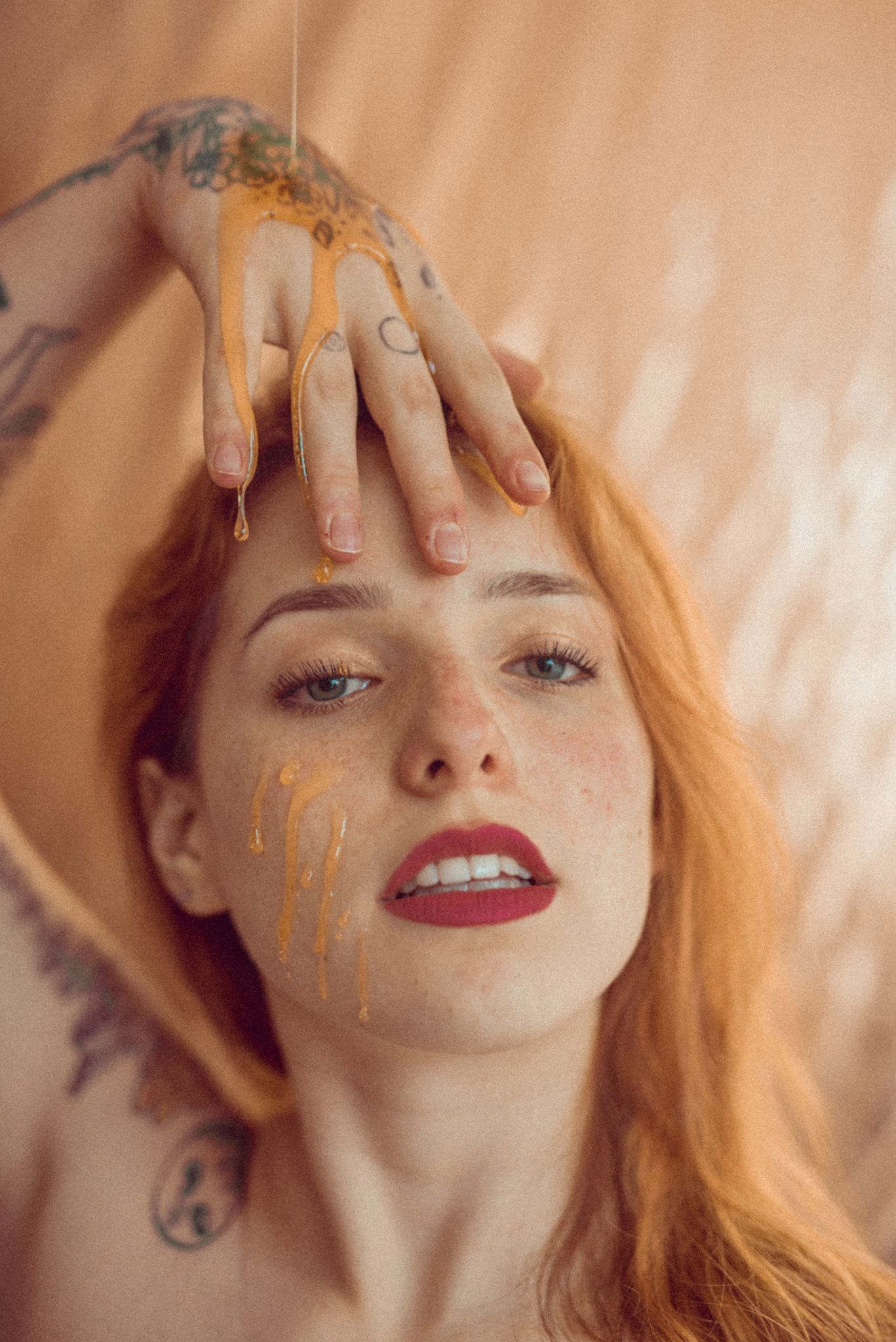 Artistic portrait featuring honey-colored paint dripping down while delicate hands rest against soft peach backdrop.
