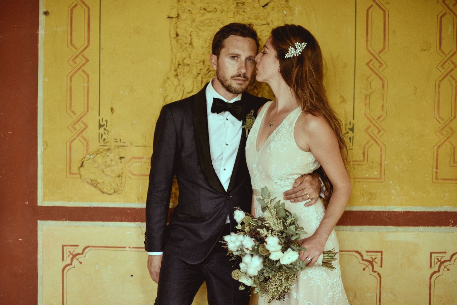 An elegant couple in formal wedding attire poses together against a gold detailed wall with a white floral bouquet.