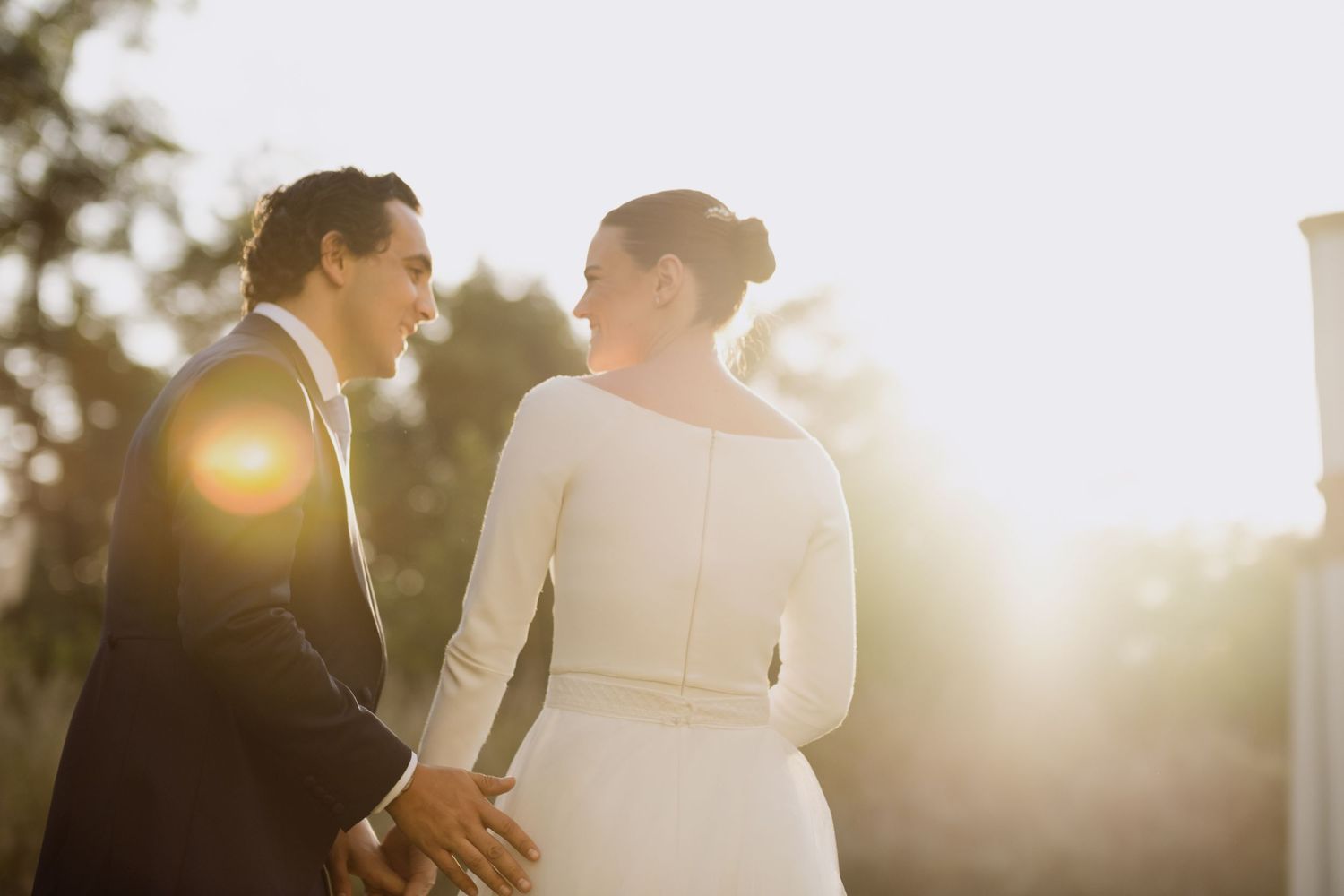 A couple in wedding attire holds hands while backlit by golden sunset light in an outdoor setting.
