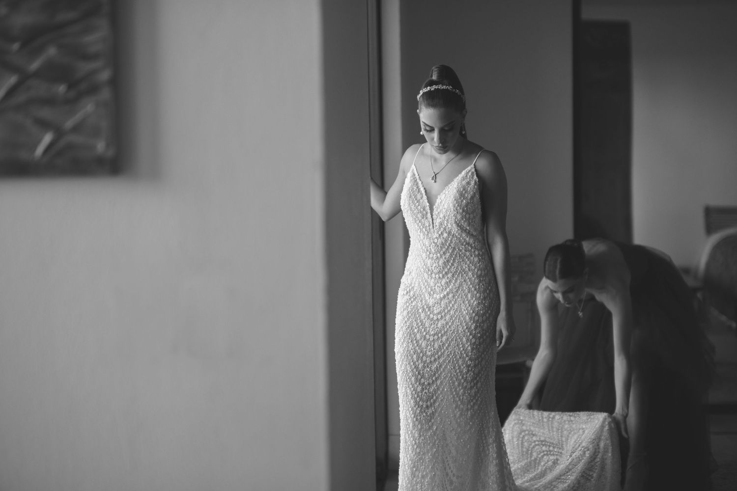 A person in a fitted lace wedding dress stands elegantly by a window in black and white photography.