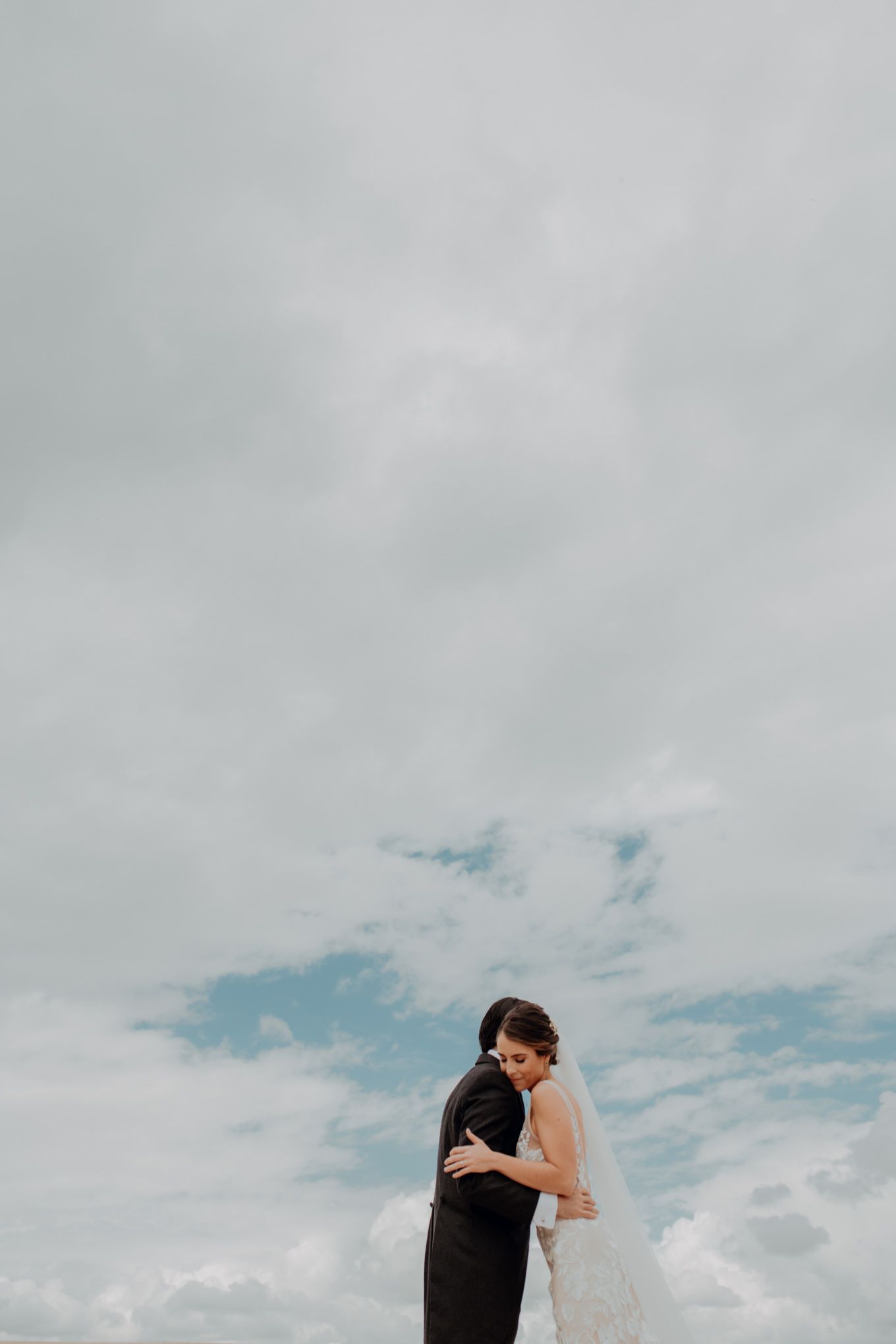 A couple embraces under cloudy skies, with the bride wearing a white dress and veil against a dramatic cloud backdrop.
