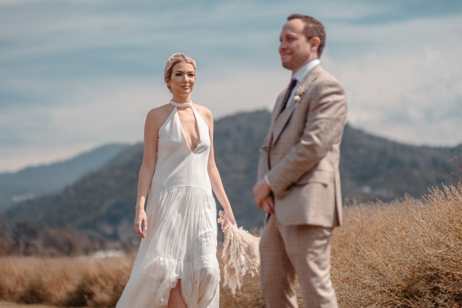 Newlyweds standing in field against mountain backdrop on sunny day.