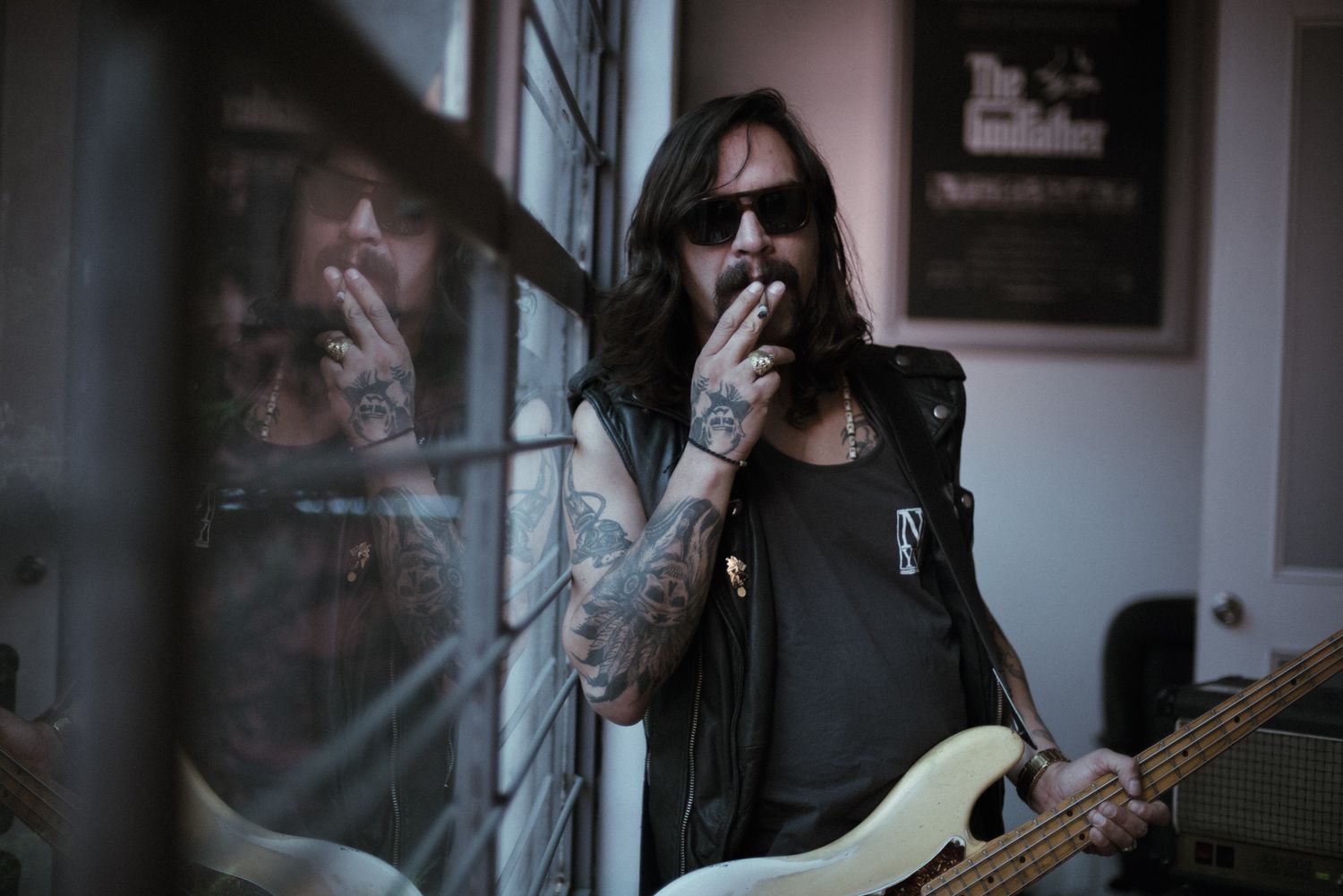 Tattooed musician in dark clothing plays guitar near window with reflective lighting.
