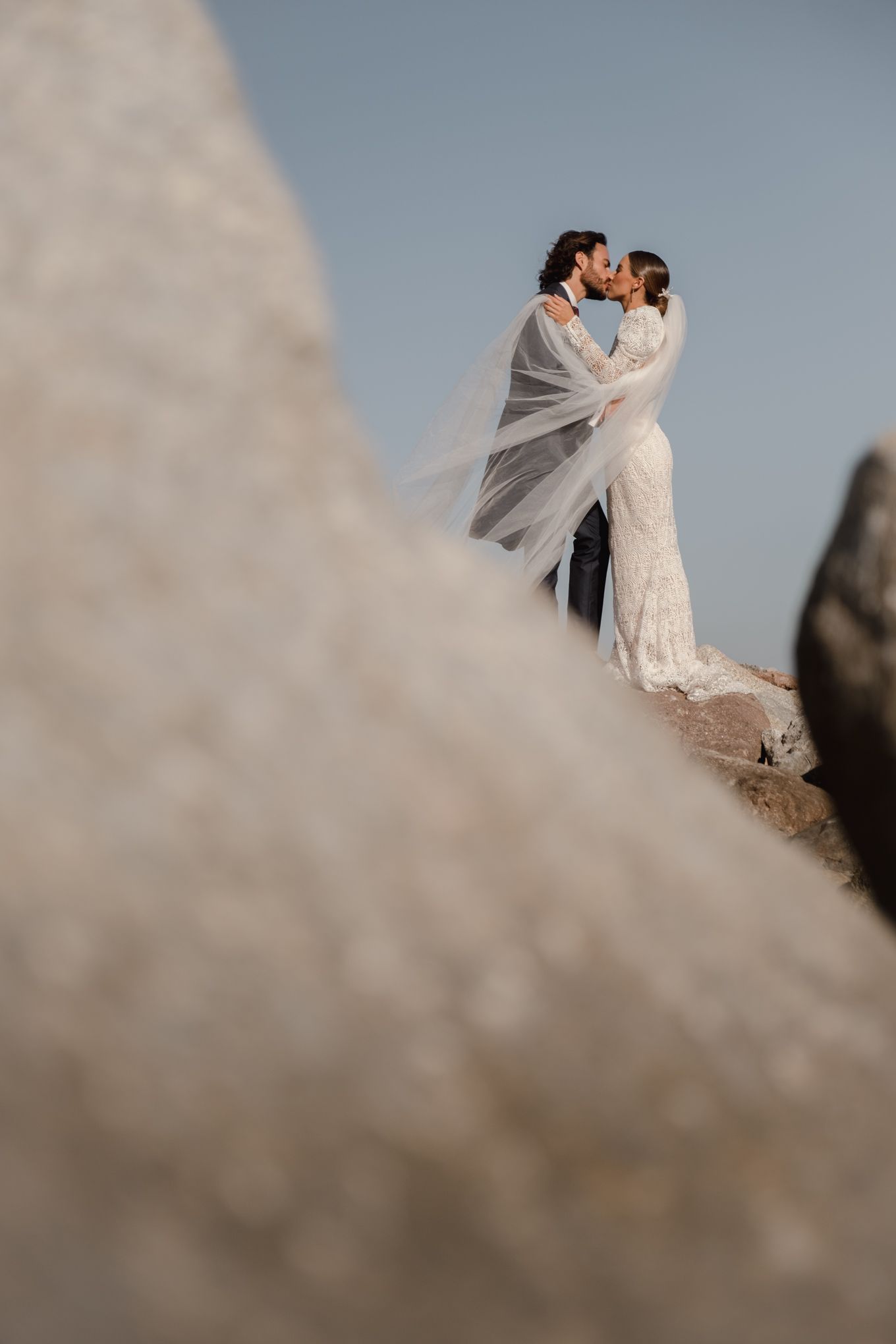 Dramatic wedding portrait against sky with flowing veil creating ethereal atmosphere.