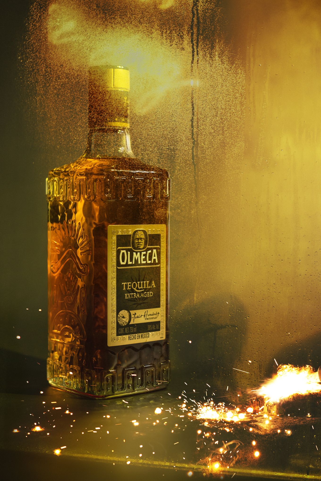 Dramatic golden-lit product shot of Olmeca tequila bottle with sparkling effects.