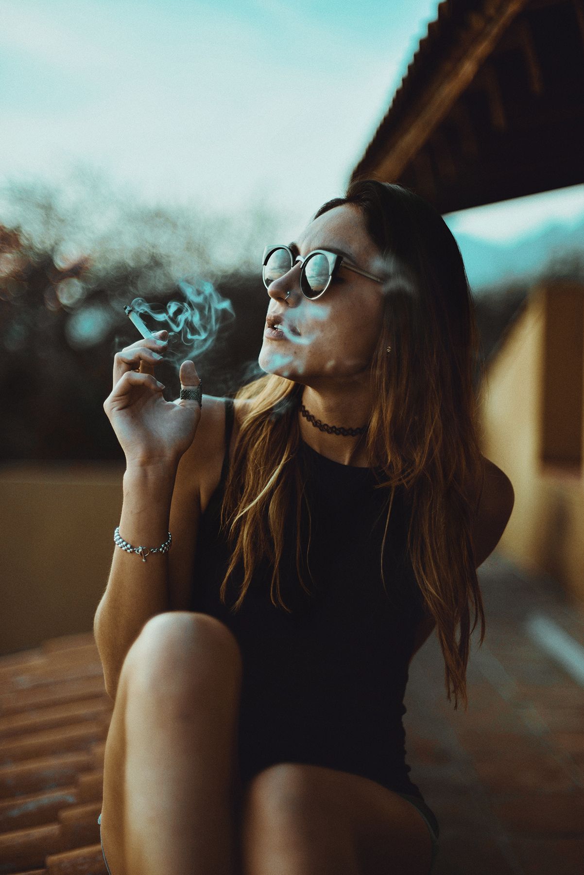 Artistic portrait of person in black bodysuit smoking on rooftop at sunset.