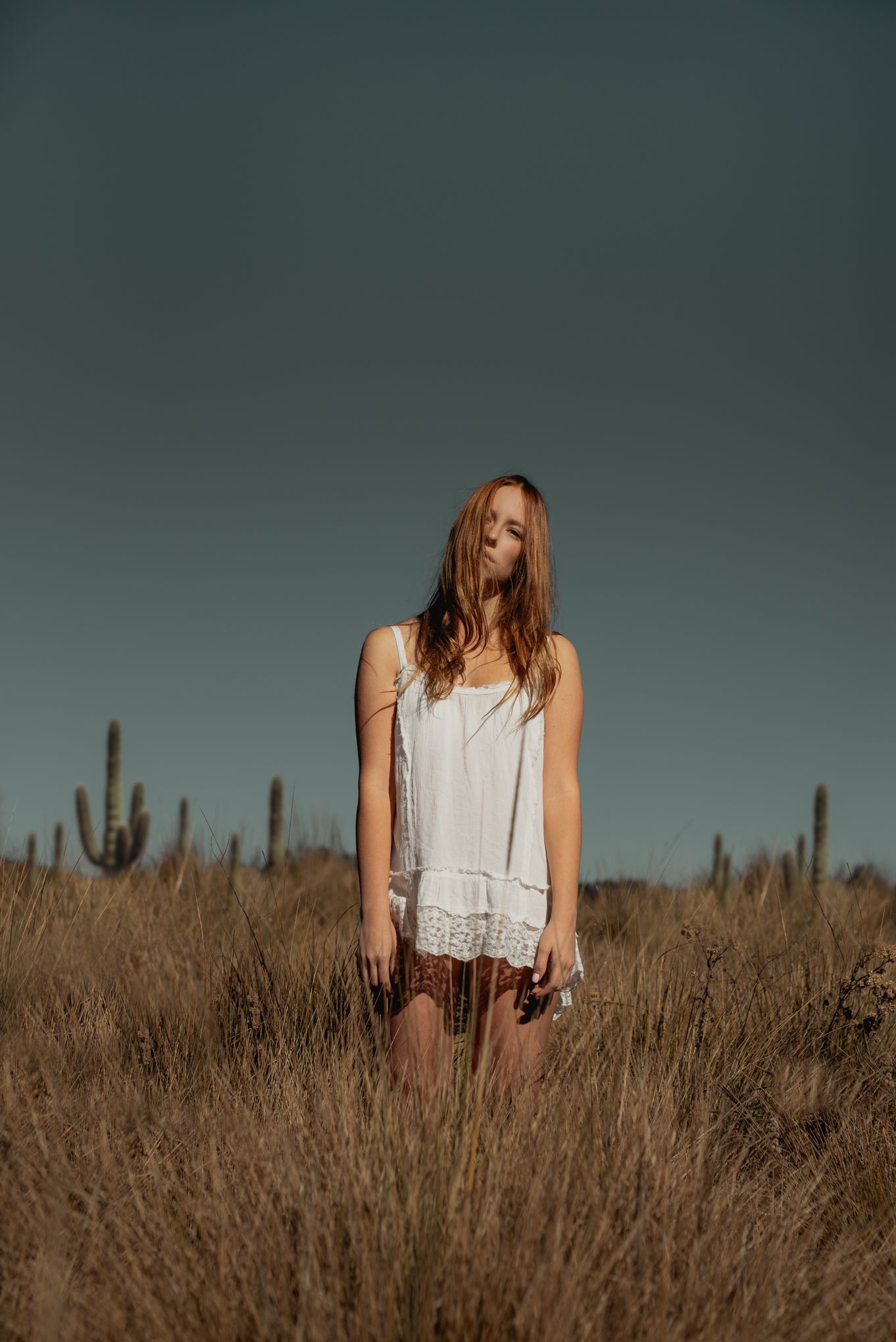 Woman in white dress stands among tall grass with cacti silhouettes against dusky sky.