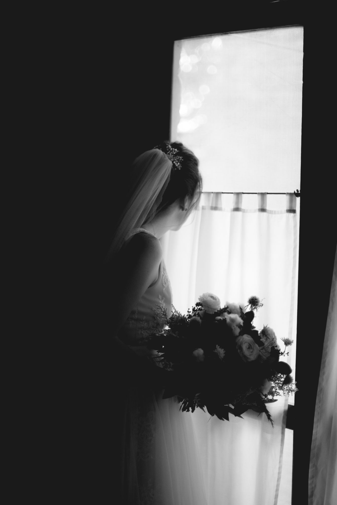 A black and white silhouette of a bride in a veil holding a bouquet while standing by a window.