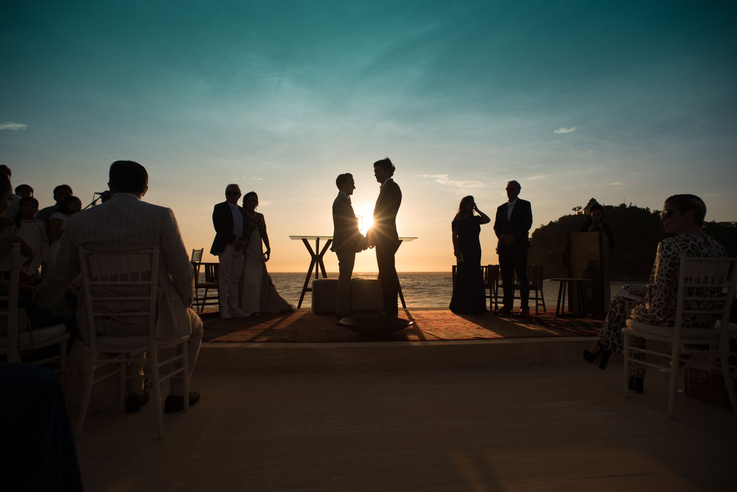 Silhouettes of people gathered at a beachfront wedding ceremony during golden sunset hour.