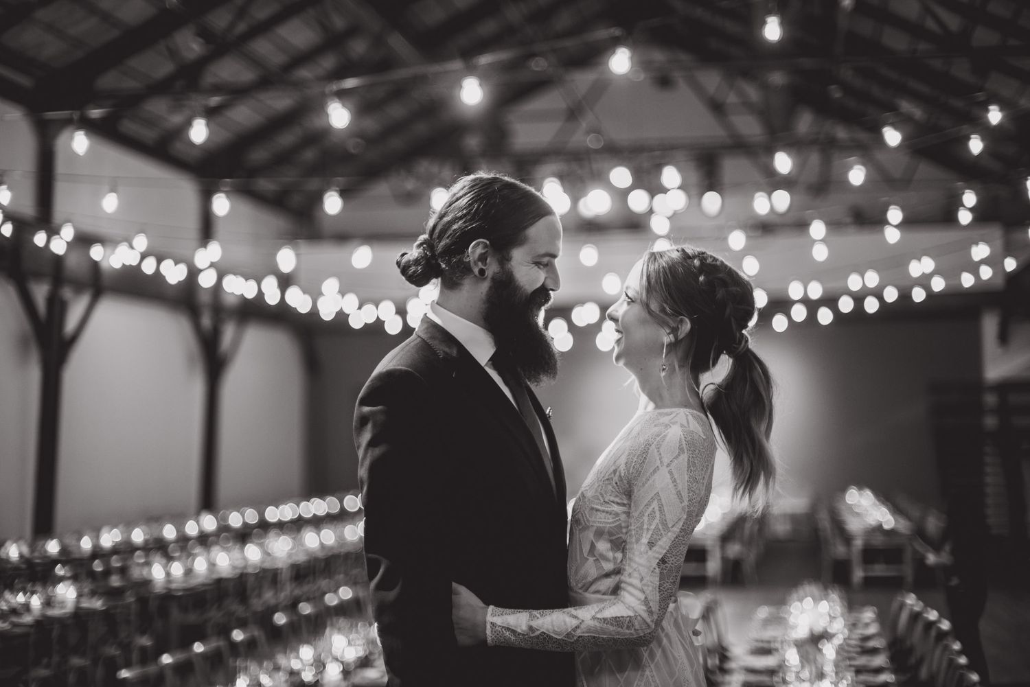 Black and white photo of a romantic moment between newlyweds dancing under twinkling string lights at their reception.