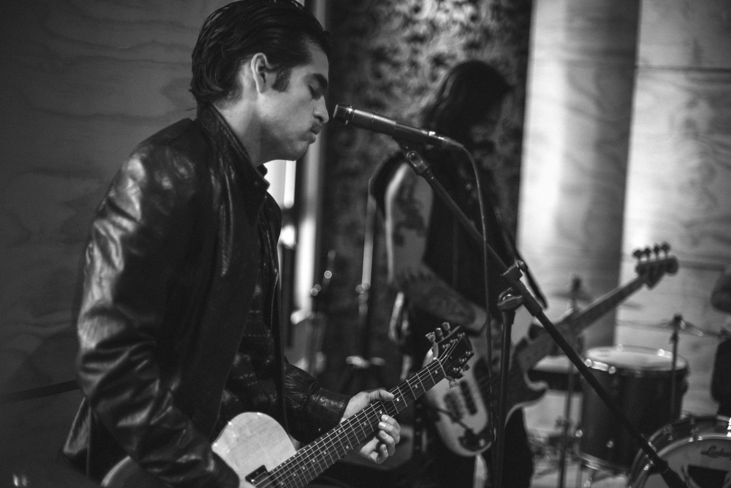 Musician performs on stage in a leather jacket with guitar and microphone in dramatic black and white lighting.