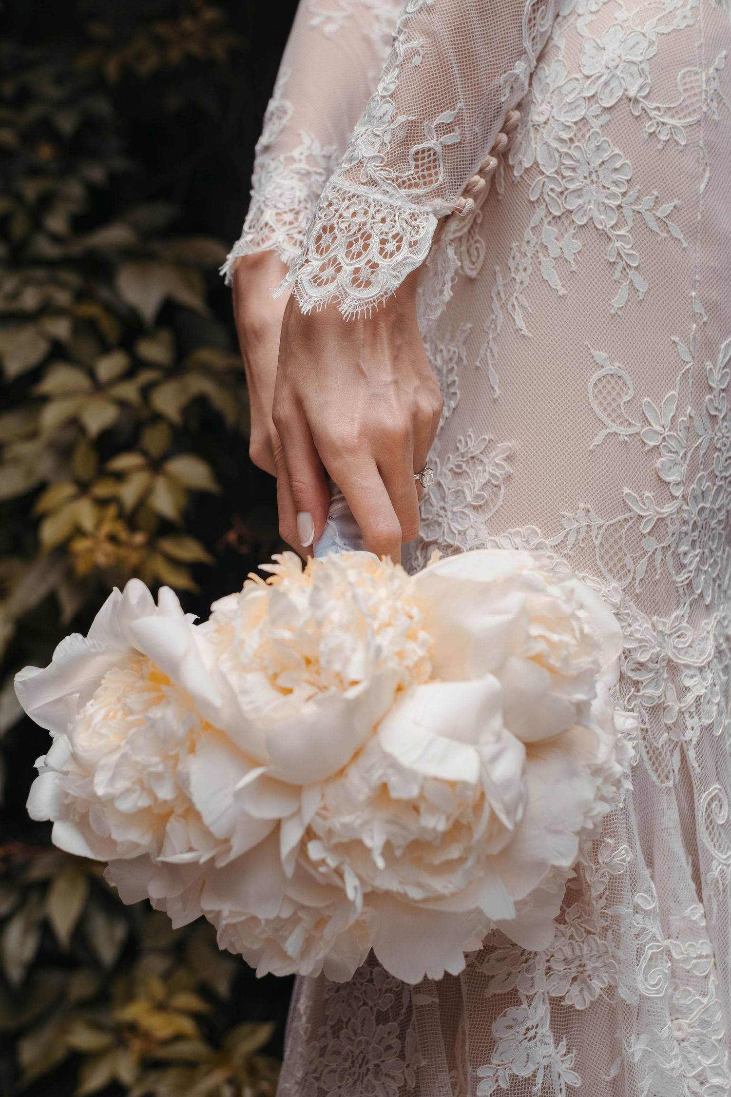 Close-up of a lace wedding dress sleeve and cream peony bouquet against dark foliage.