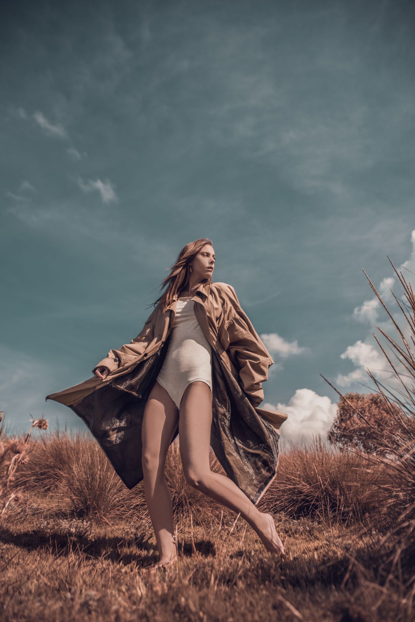 Artistic fashion shot in brown coat and bodysuit against dramatic stormy sky.