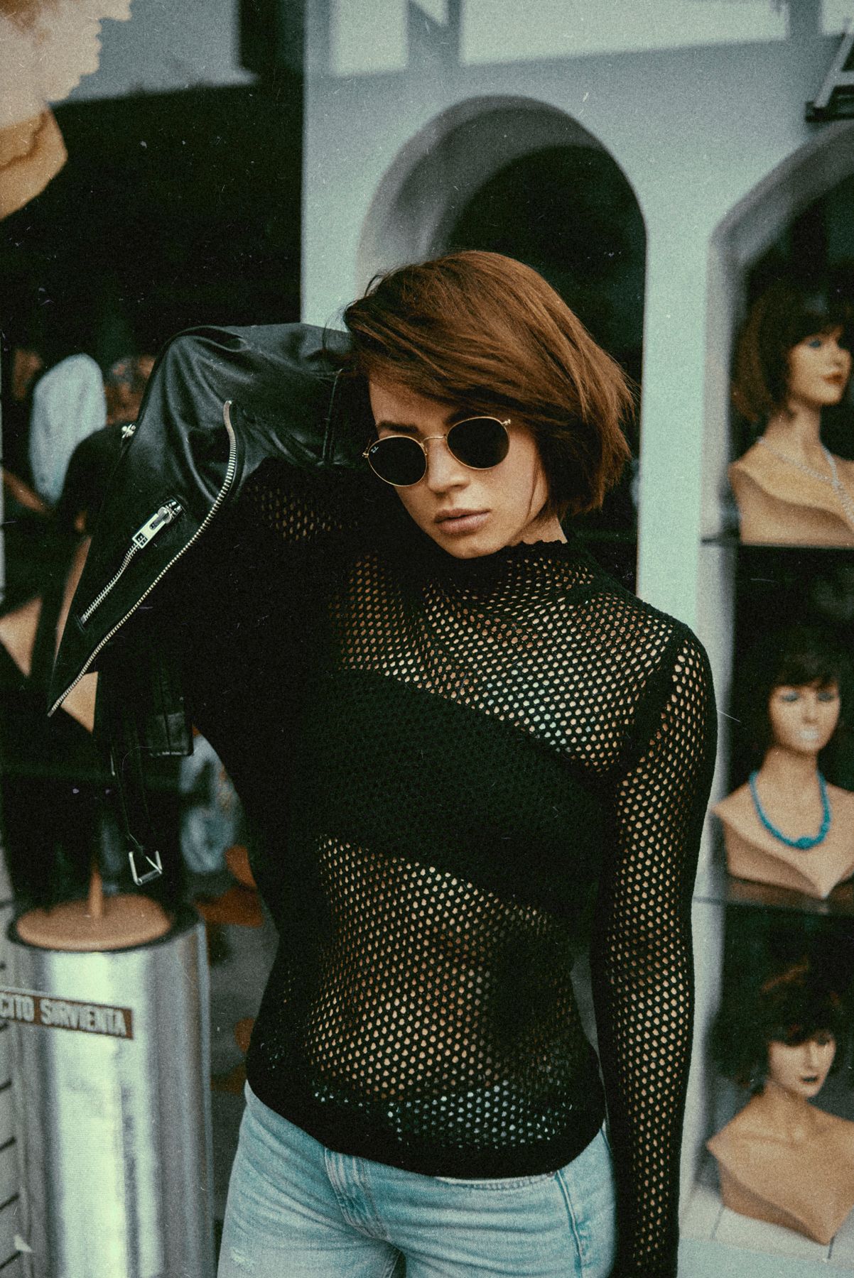 Fashion portrait with mesh top, leather jacket and sunglasses against mannequin display.