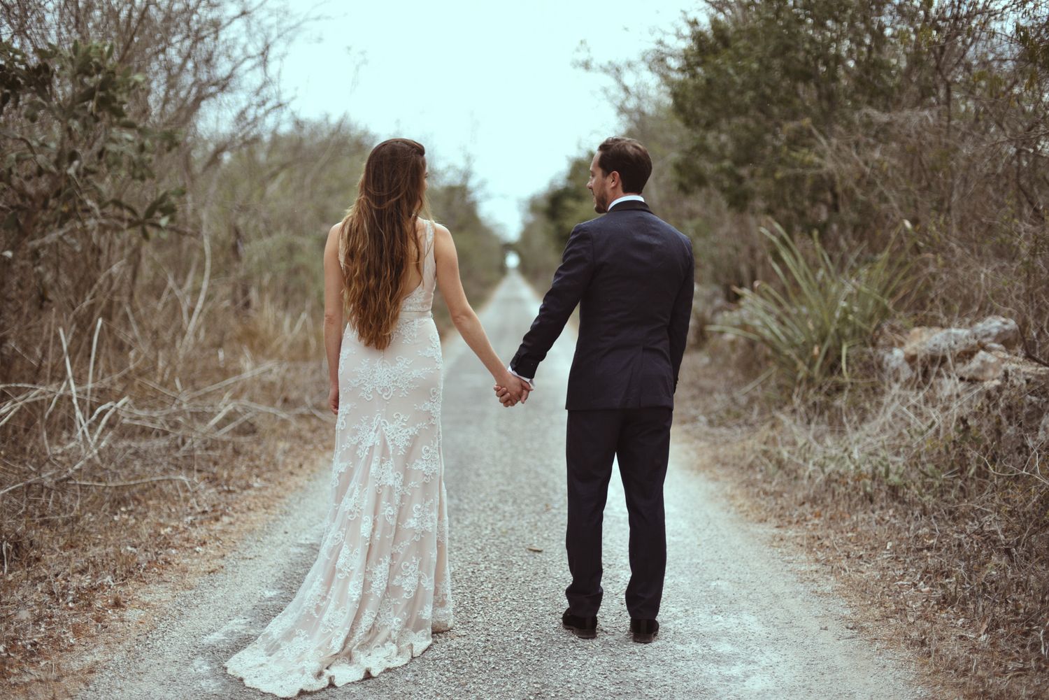 A couple in wedding attire walking hand in hand down a rustic dirt path surrounded by shrubs and trees.