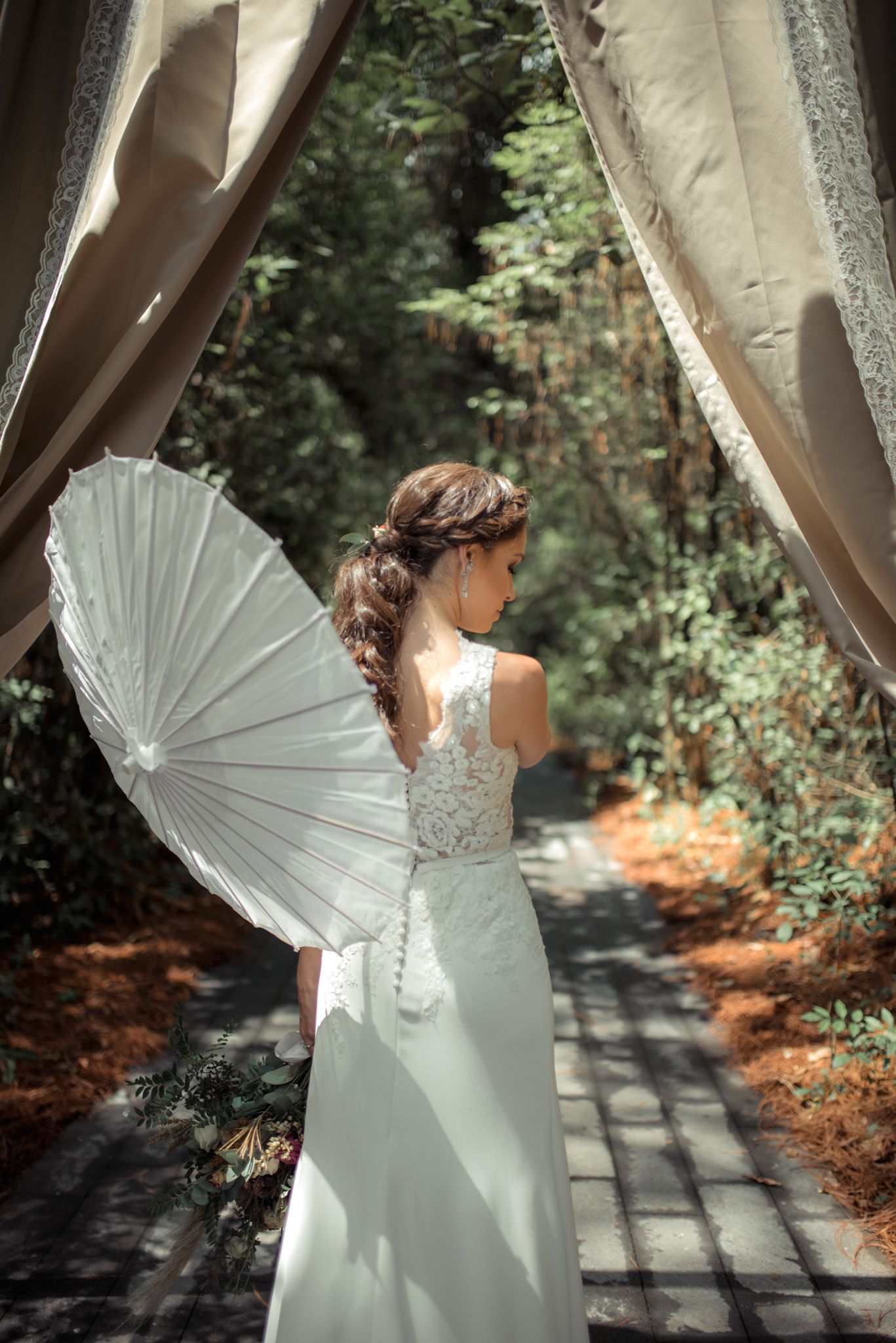 An ethereal bridal portrait from behind shows a white parasol and elegant lace gown on a forest path framed by curtains.