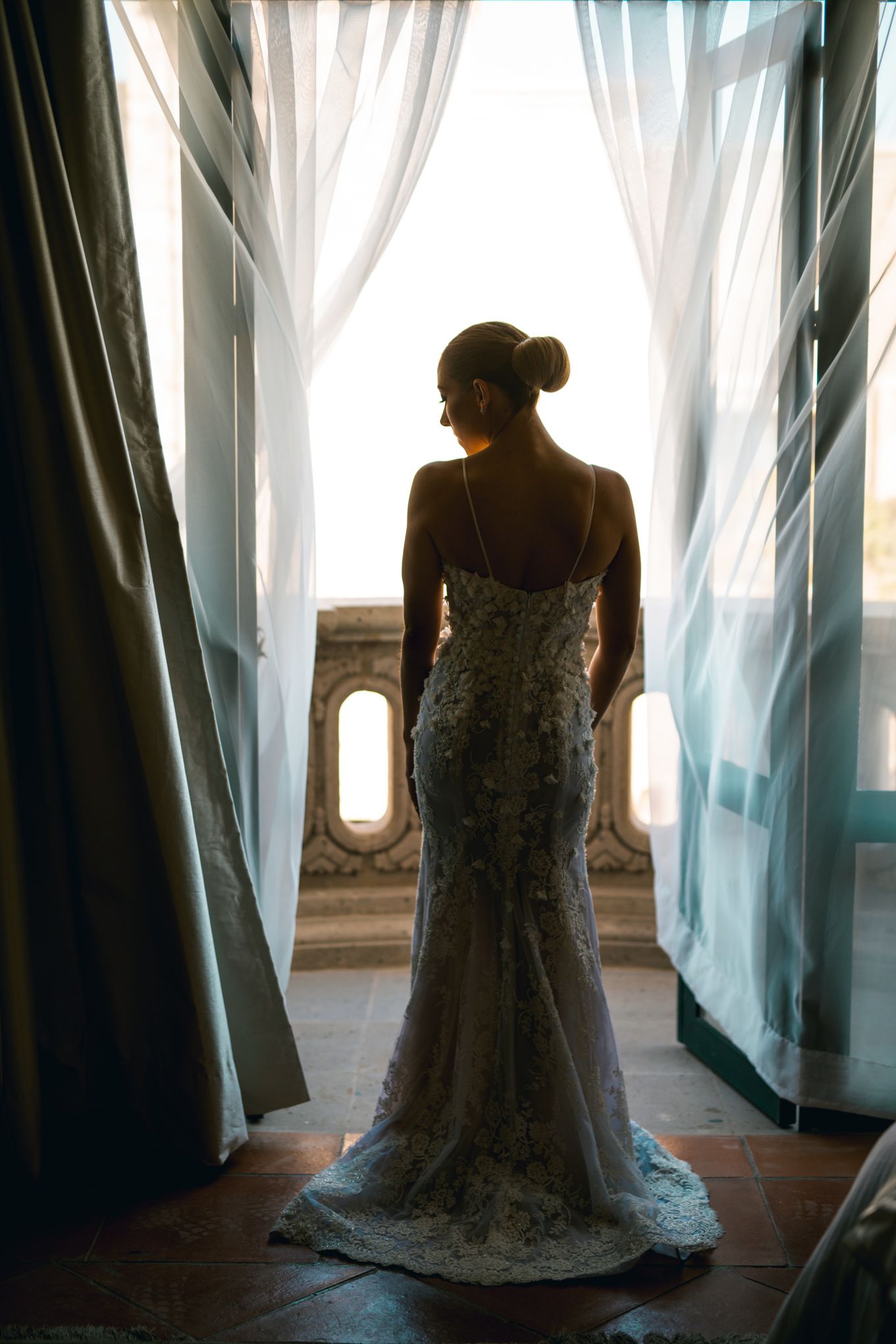 A silhouetted figure in a lace wedding dress stands elegantly by sunlit windows, creating a romantic backlit effect.