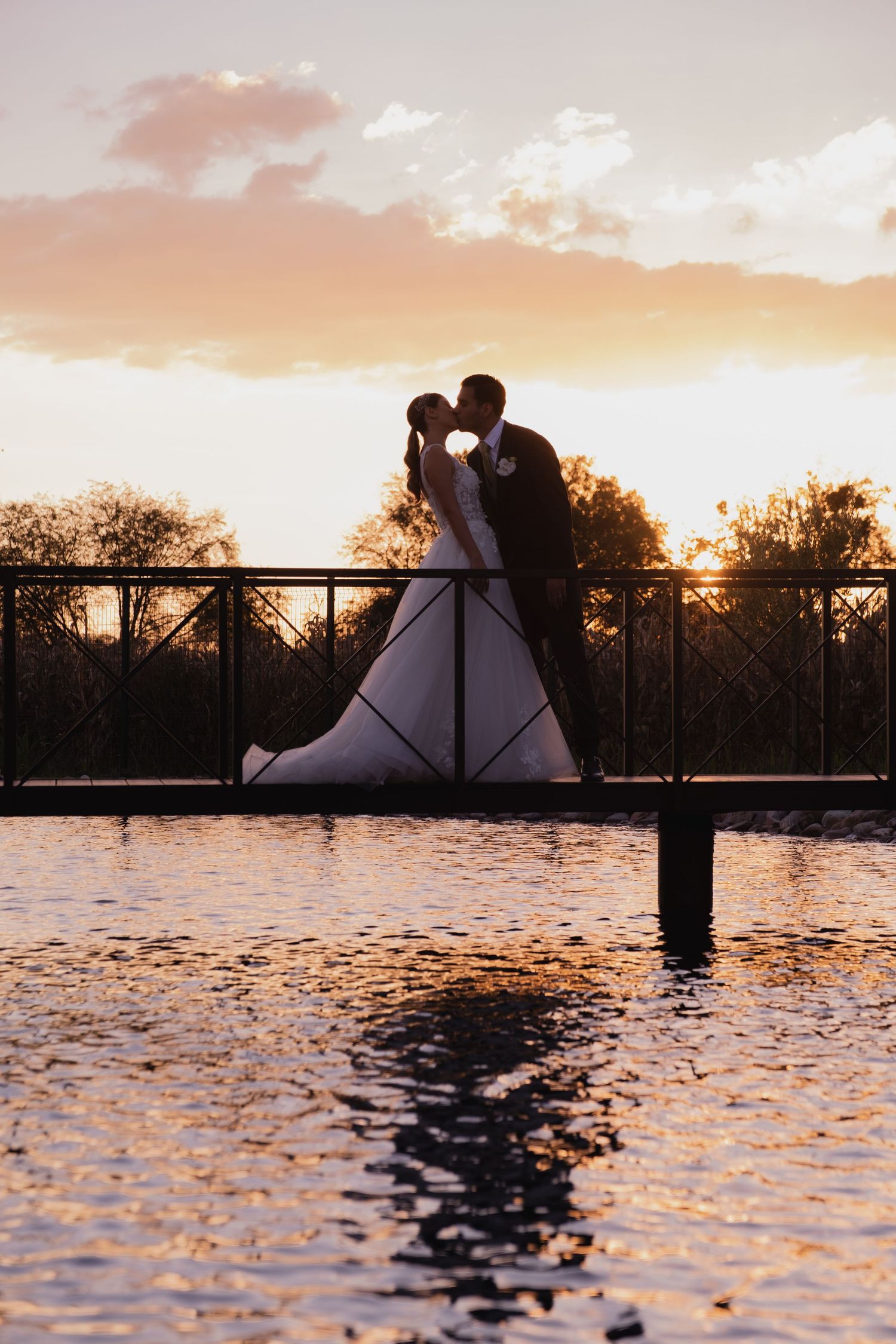 A romantic silhouette of a bride and groom embrace on a bridge during sunset with golden sky and water reflections.