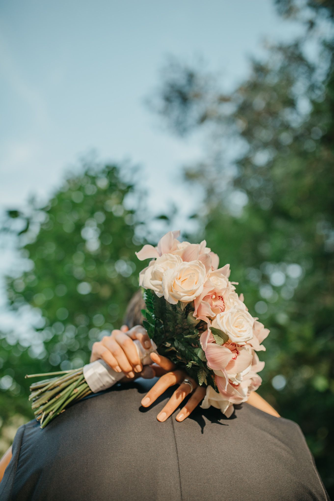A peach rose wedding bouquet held over a person wearing a gray suit, photographed from behind against blurred greenery.