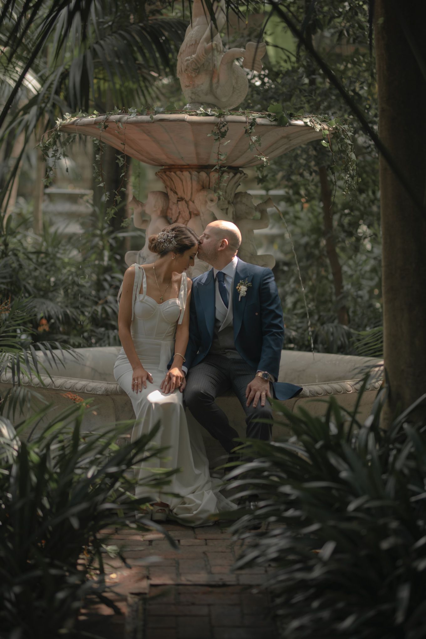 A romantic couple shares a moment together by a garden fountain surrounded by lush greenery.