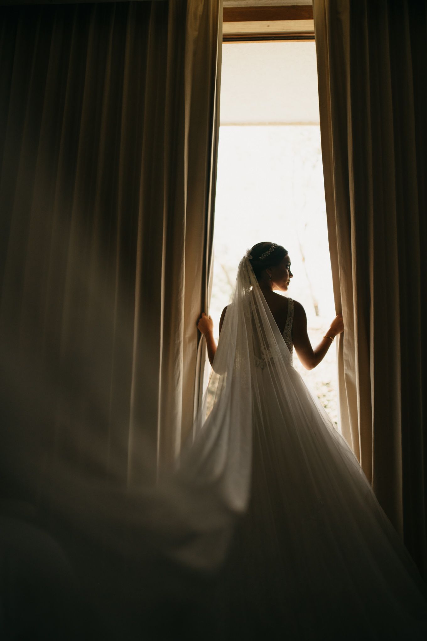 A silhouetted figure in a flowing wedding veil stands between dark curtains against a bright window backlight.