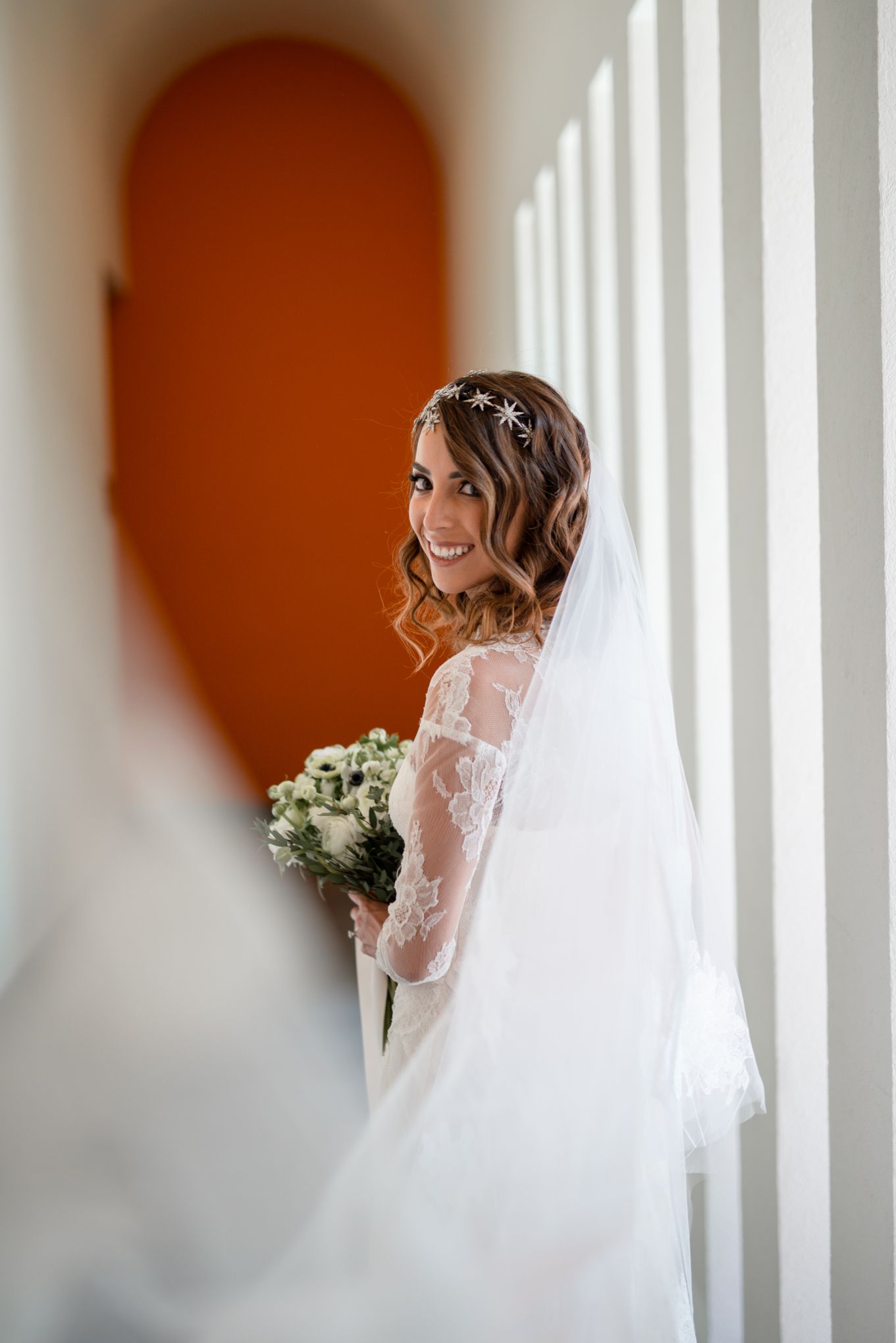 A bride in a white lace gown with long sleeves and flowing veil smiles while holding a white floral bouquet.