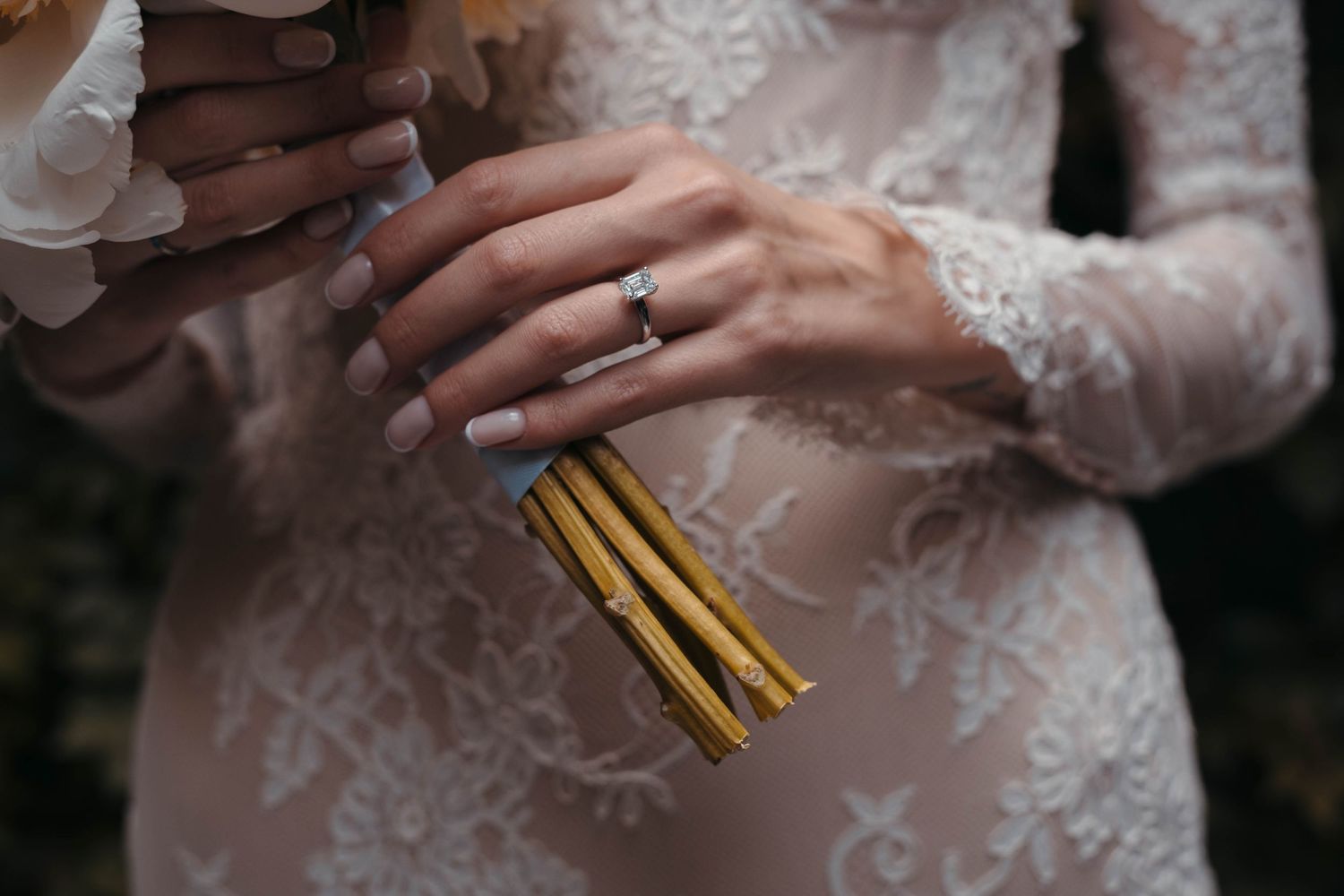 Delicate lace detail of a wedding dress with engagement ring and gold pins visible.