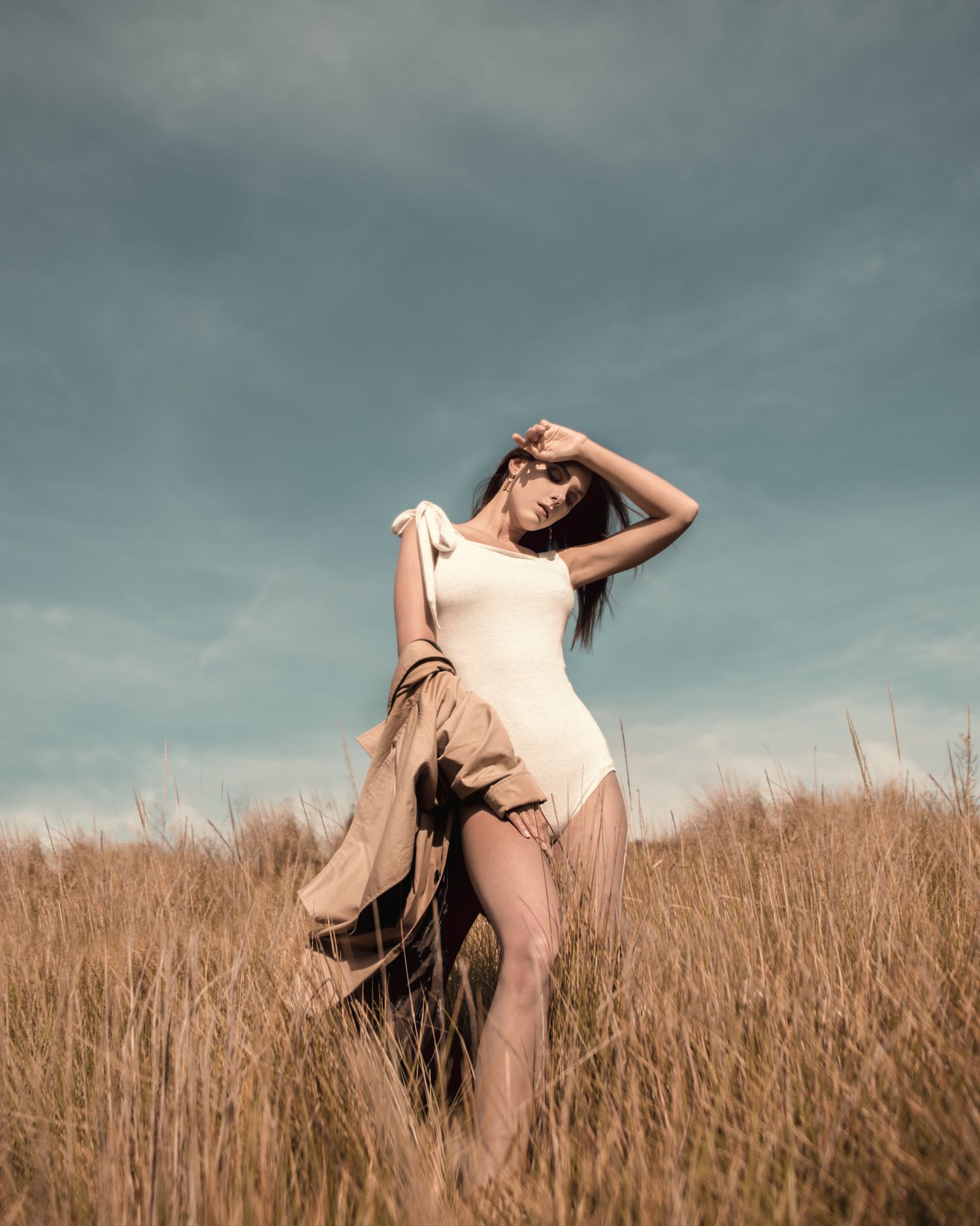 Ethereal figure in flowing dress poses dramatically in tall grass field under cloudy sky.